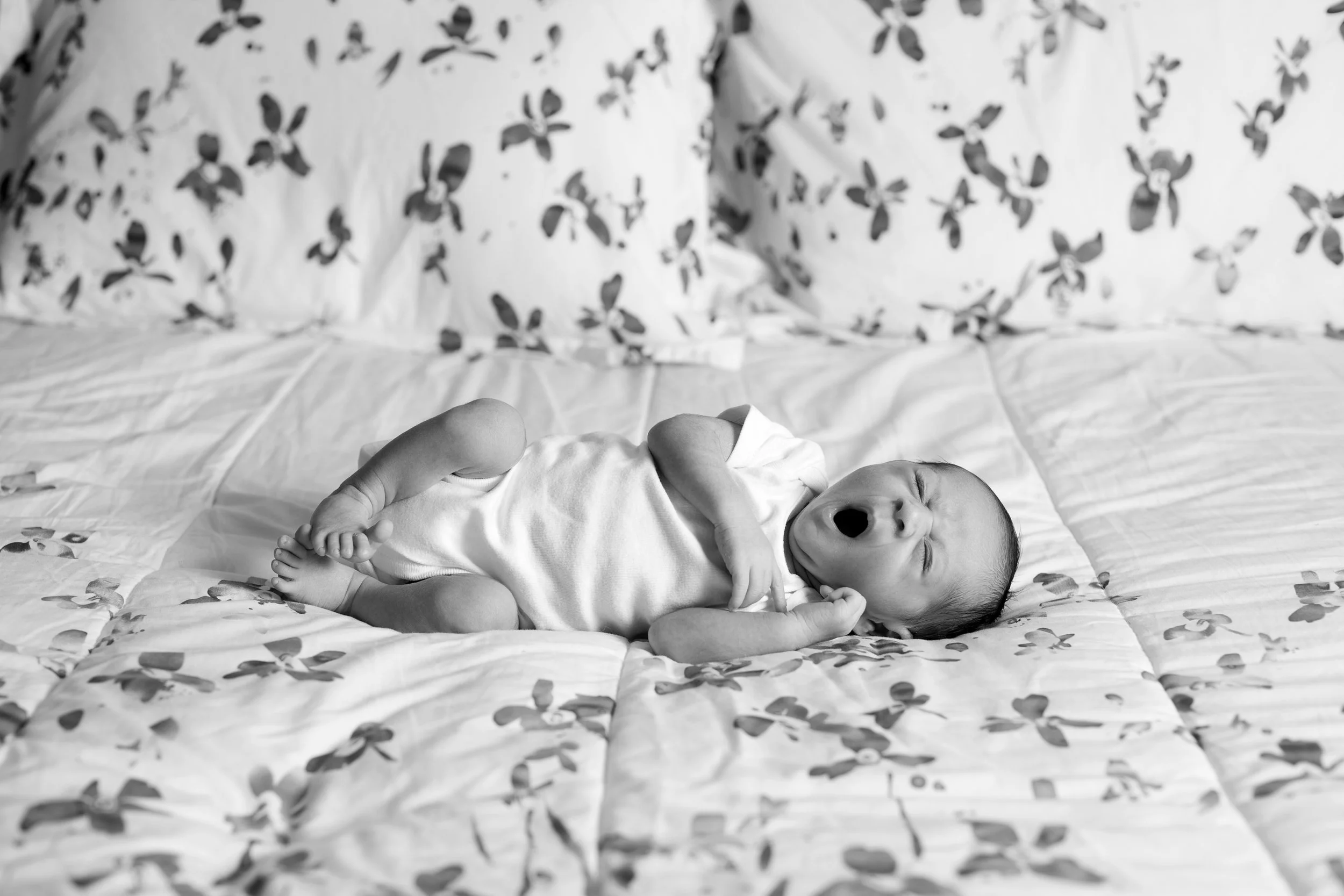 A baby yawning while lying on a bed with patterned sheets and pillowcases.
