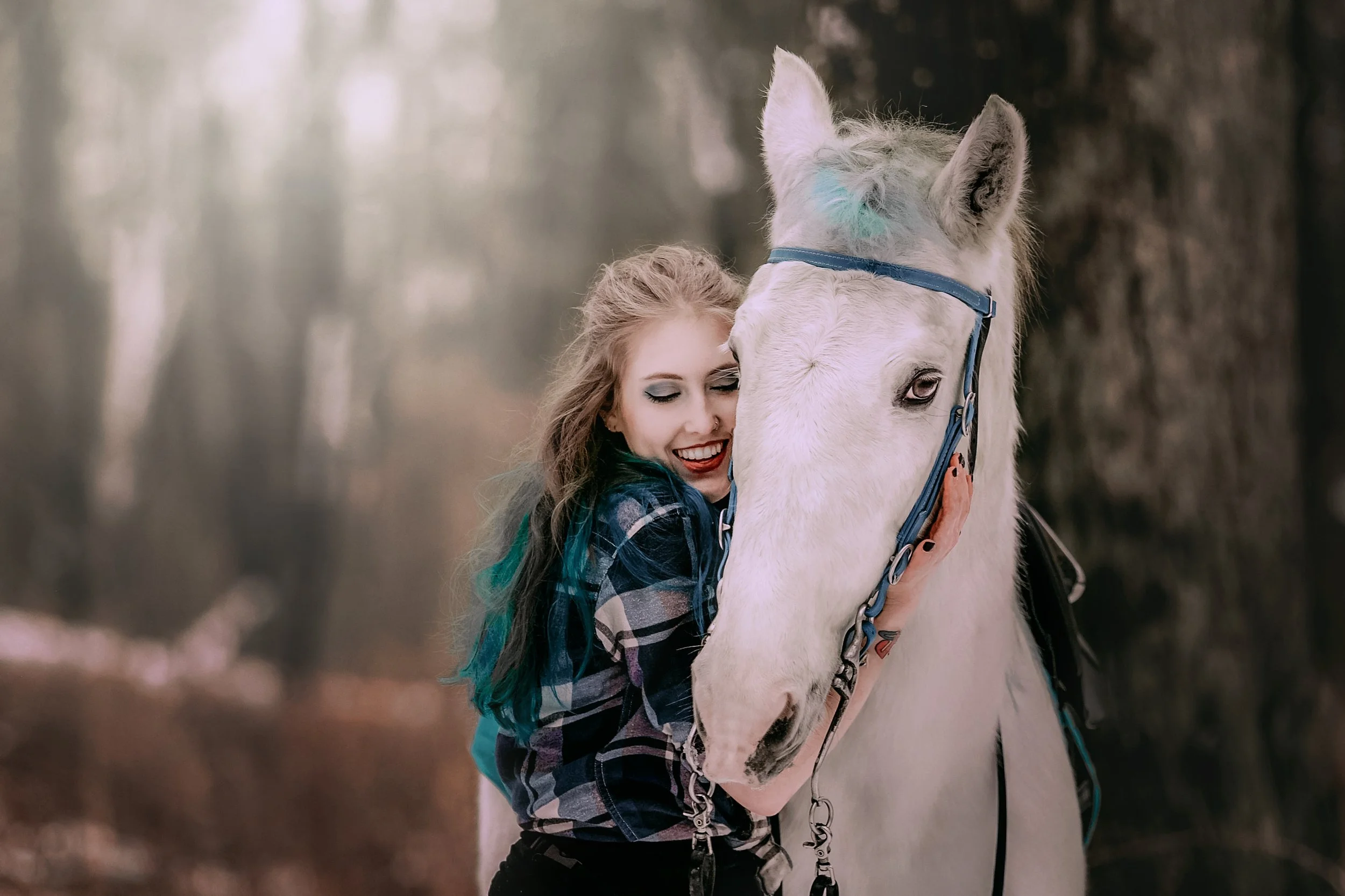 A young woman in a plaid shirt hugging a white horse in a wooded outdoor setting.