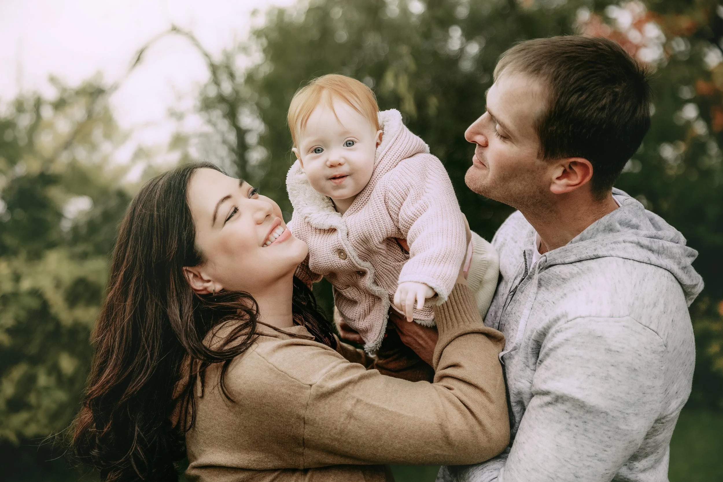 A happy family of three outdoors, with a woman holding a toddler girl and a man standing beside them, smiling at each other.