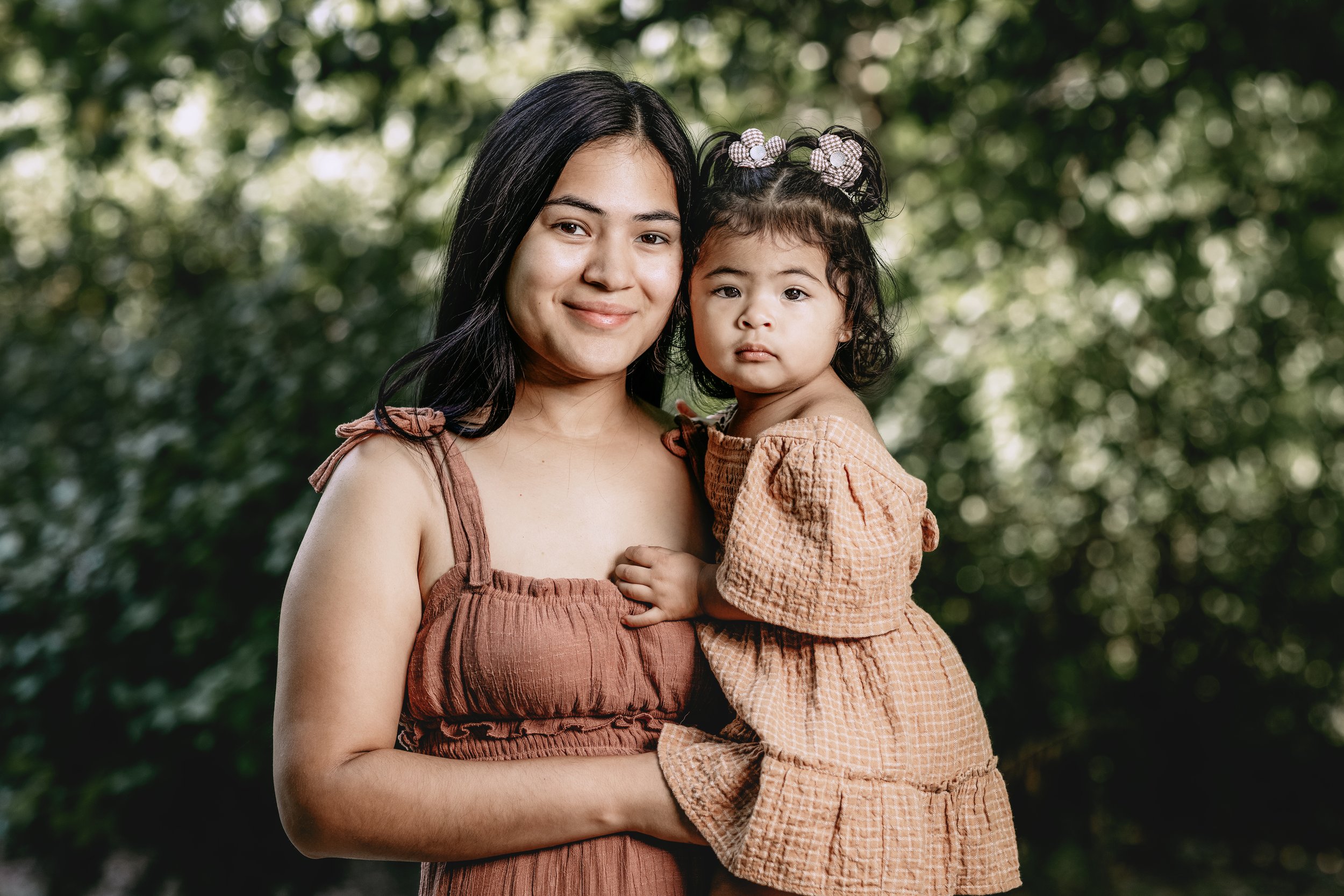 A woman holding a young girl outdoors, both wearing brown dresses, with green foliage background.