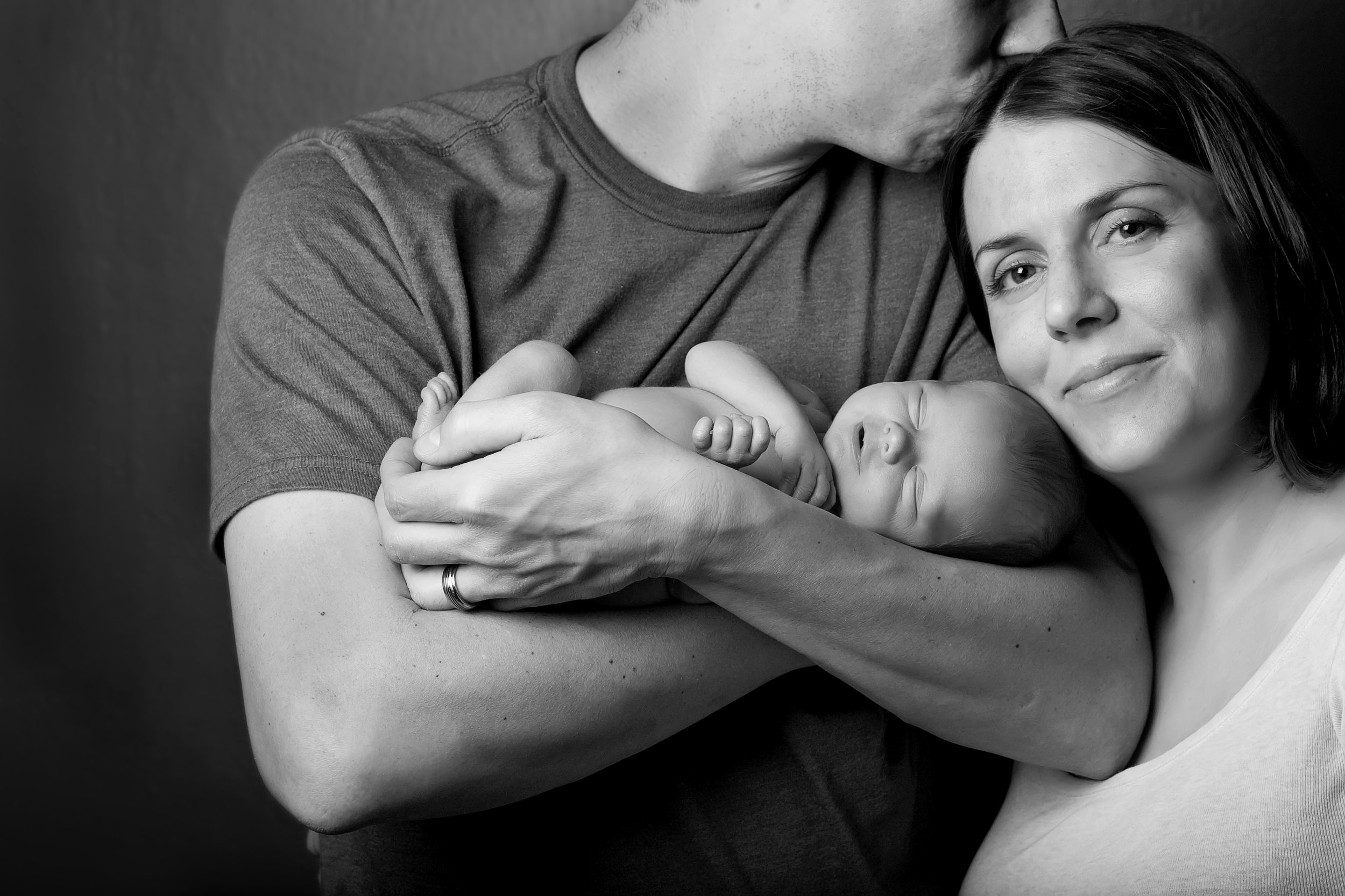 A black-and-white photo of a woman holding a sleeping baby, with a man leaning in and kissing the baby's head, all smiling softly.