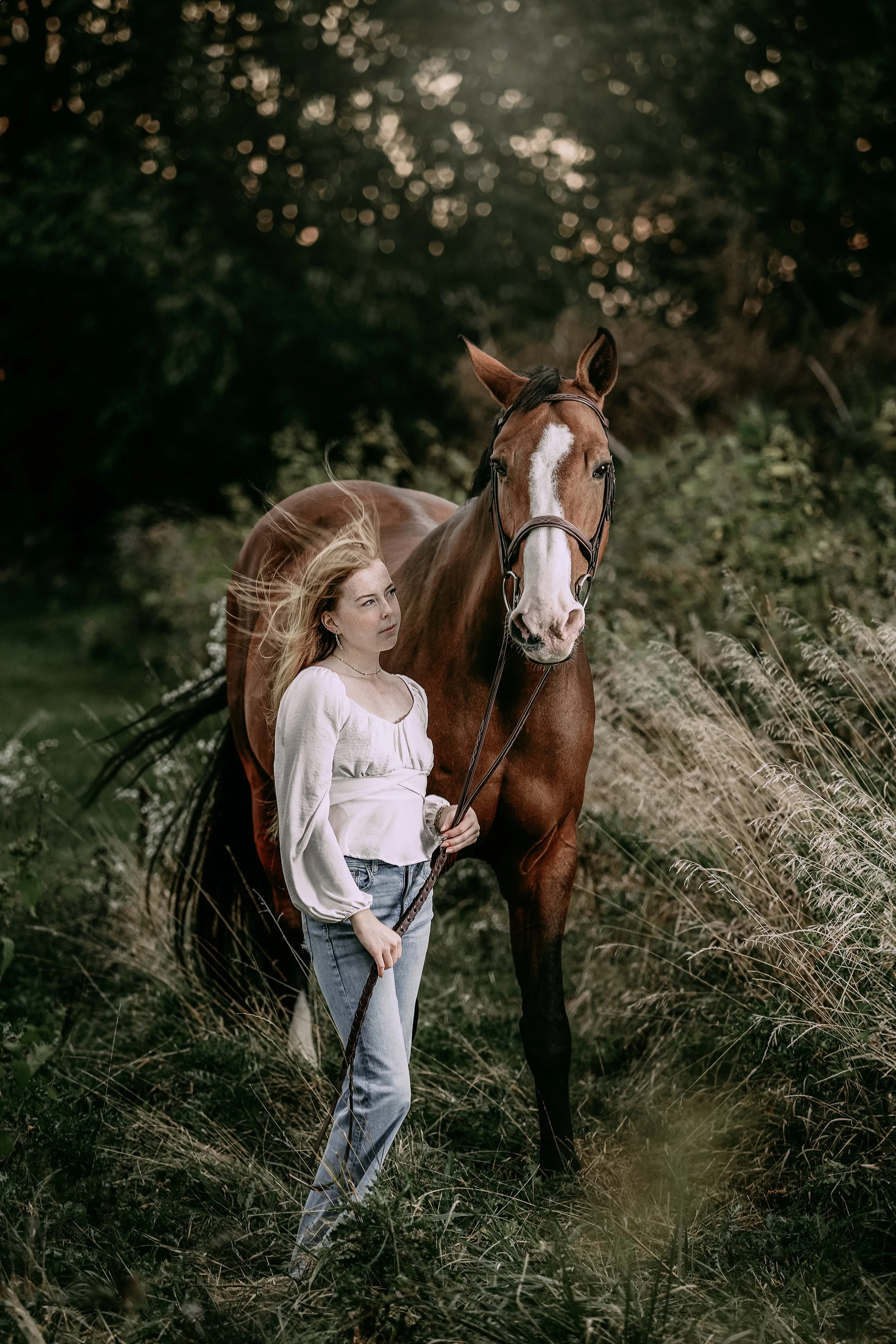 A young woman with long blonde hair standing outdoors in a grassy area, holding a lead rope attached to a chestnut horse, with trees and foliage in the background.