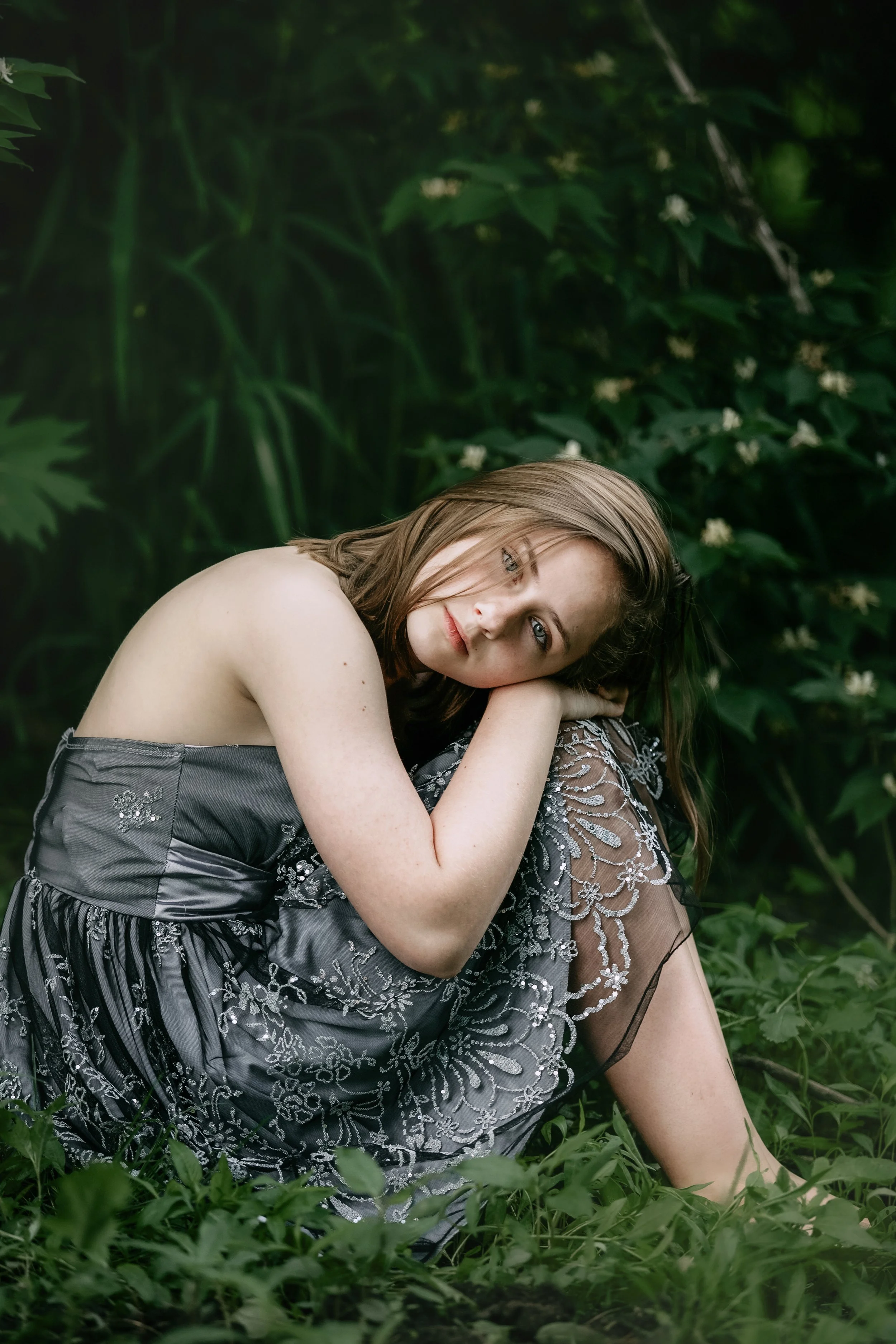A young woman with brown hair and blue eyes sits in a green outdoor setting, resting her head on her knees and looking at the camera.
