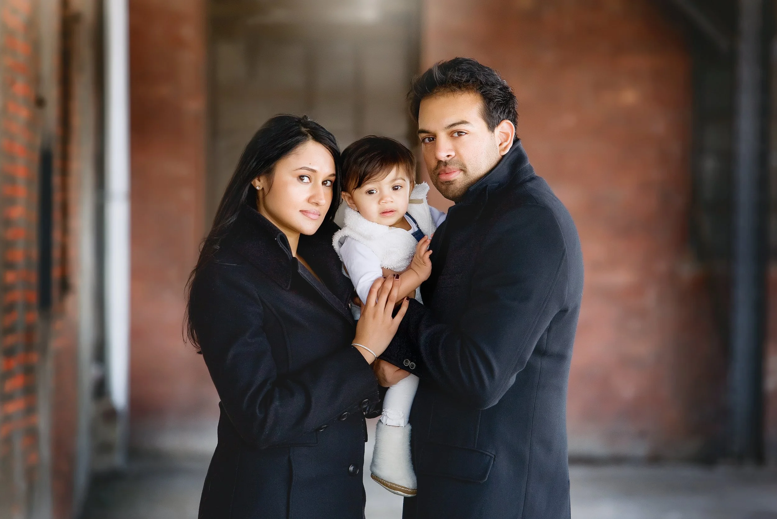 Portrait of a young ethnic couple with their toddler girl outdoors in an urban setting with brick walls.