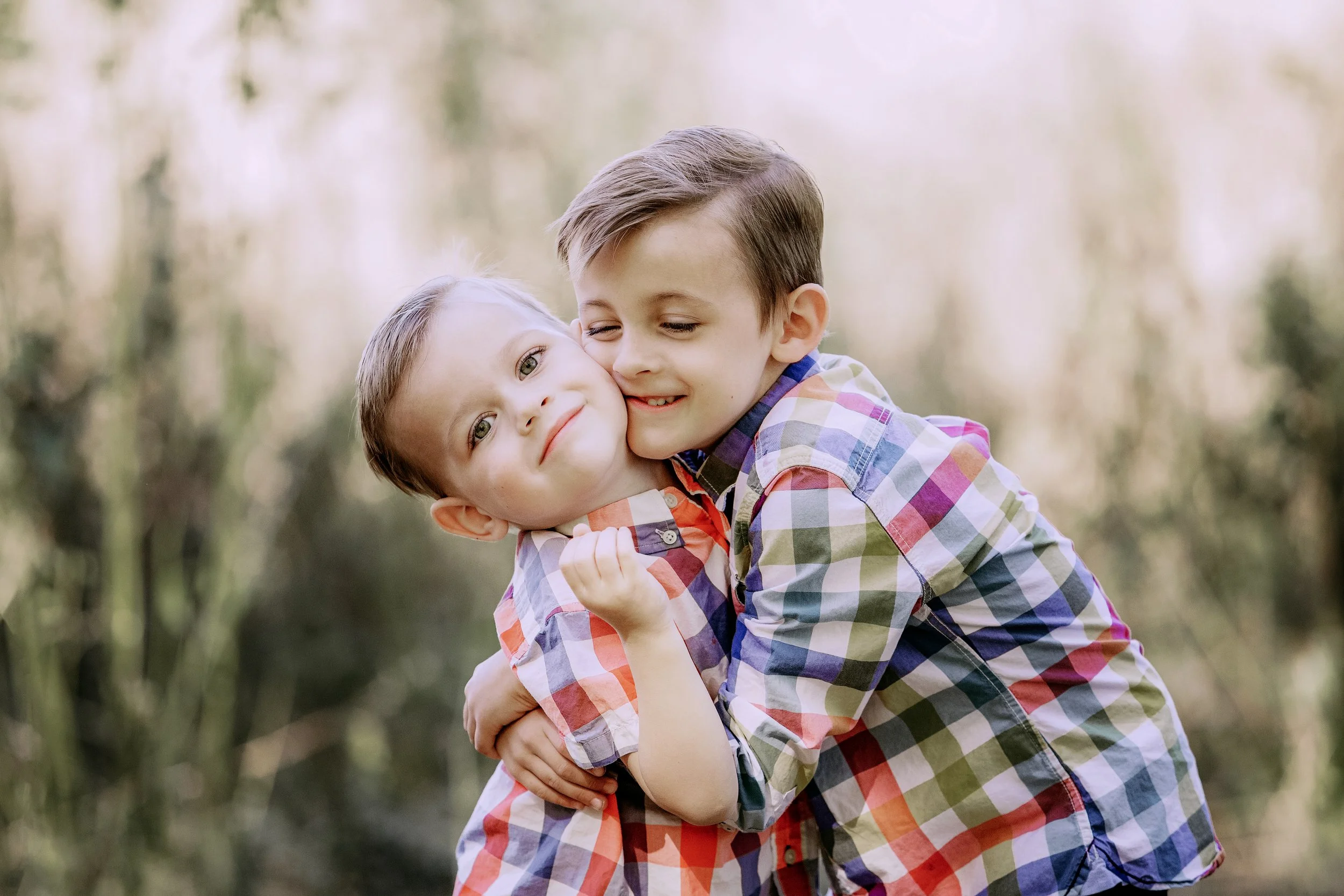 Two young boys hugging outdoors in a park or forest with blurred trees in the background.