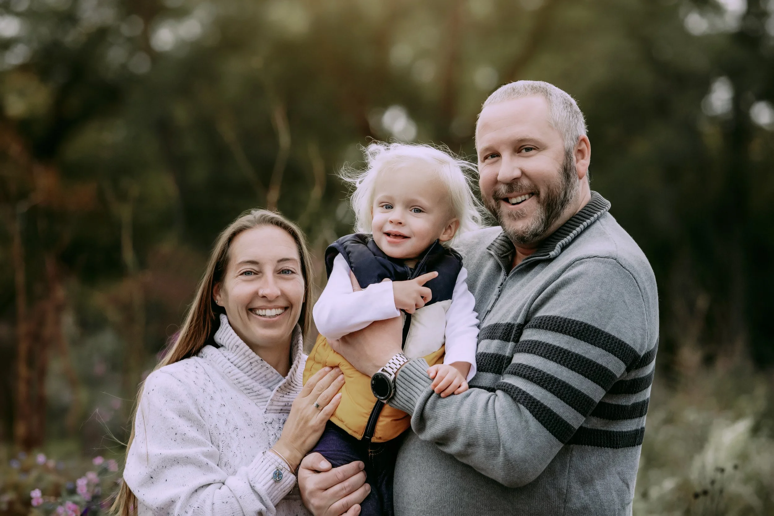 A happy family of three outdoors during fall, consisting of a smiling woman with long hair, a man with a beard, and a young girl with blonde hair, all enjoying a moment together.