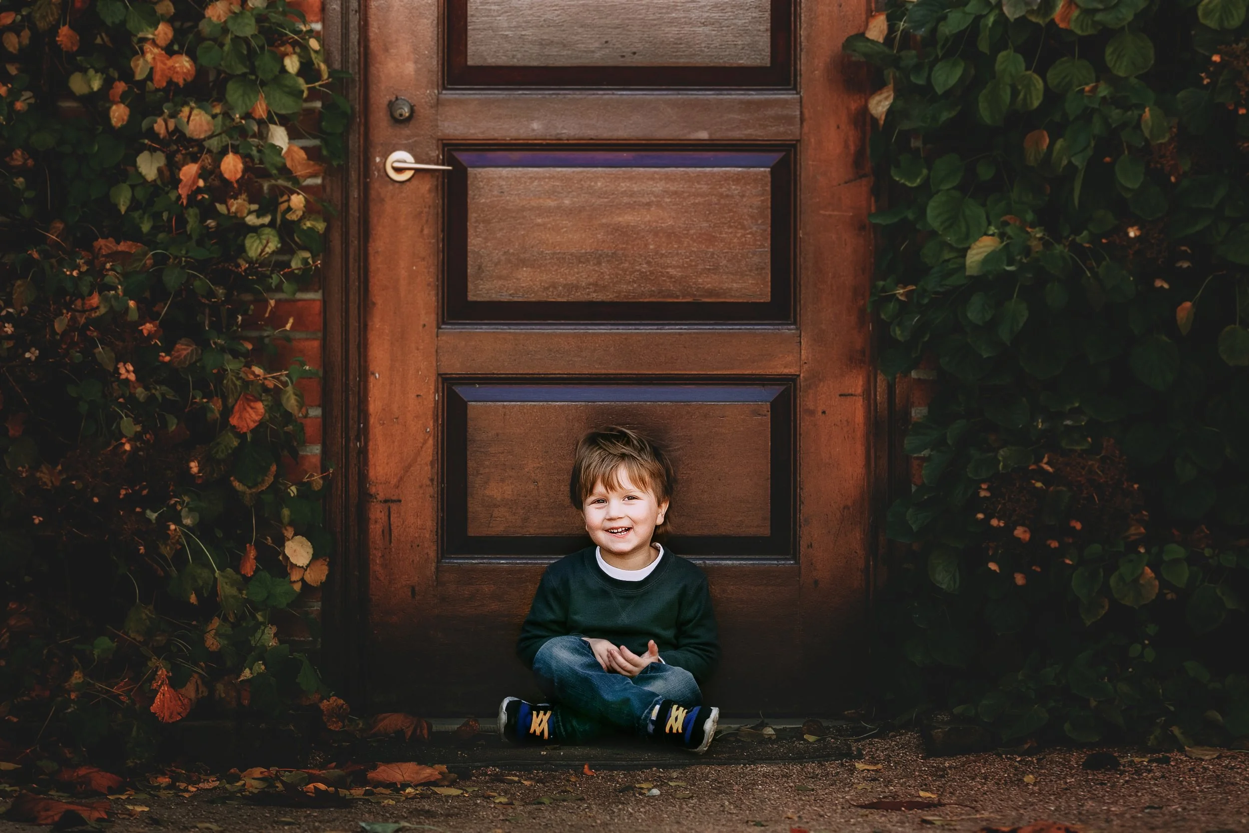 A young boy with brown hair, wearing a dark sweater and jeans, sitting on the ground in front of a wooden door, smiling.