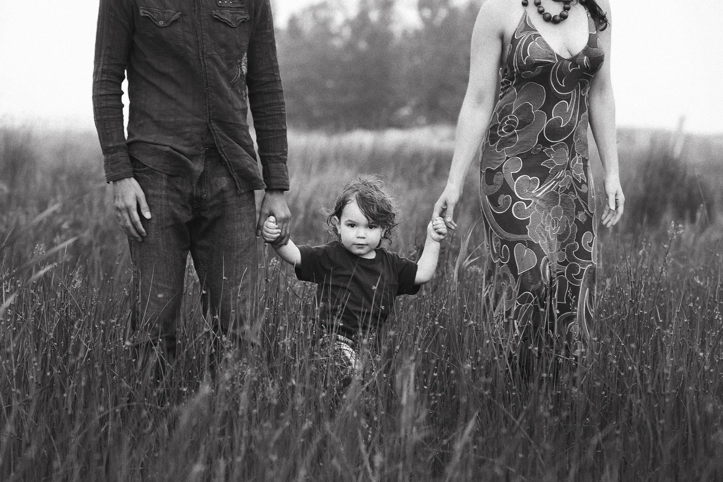 A family walking through a grassy field holding their child's hands, with only the lower half of two adults visible and the child looking directly at the camera.