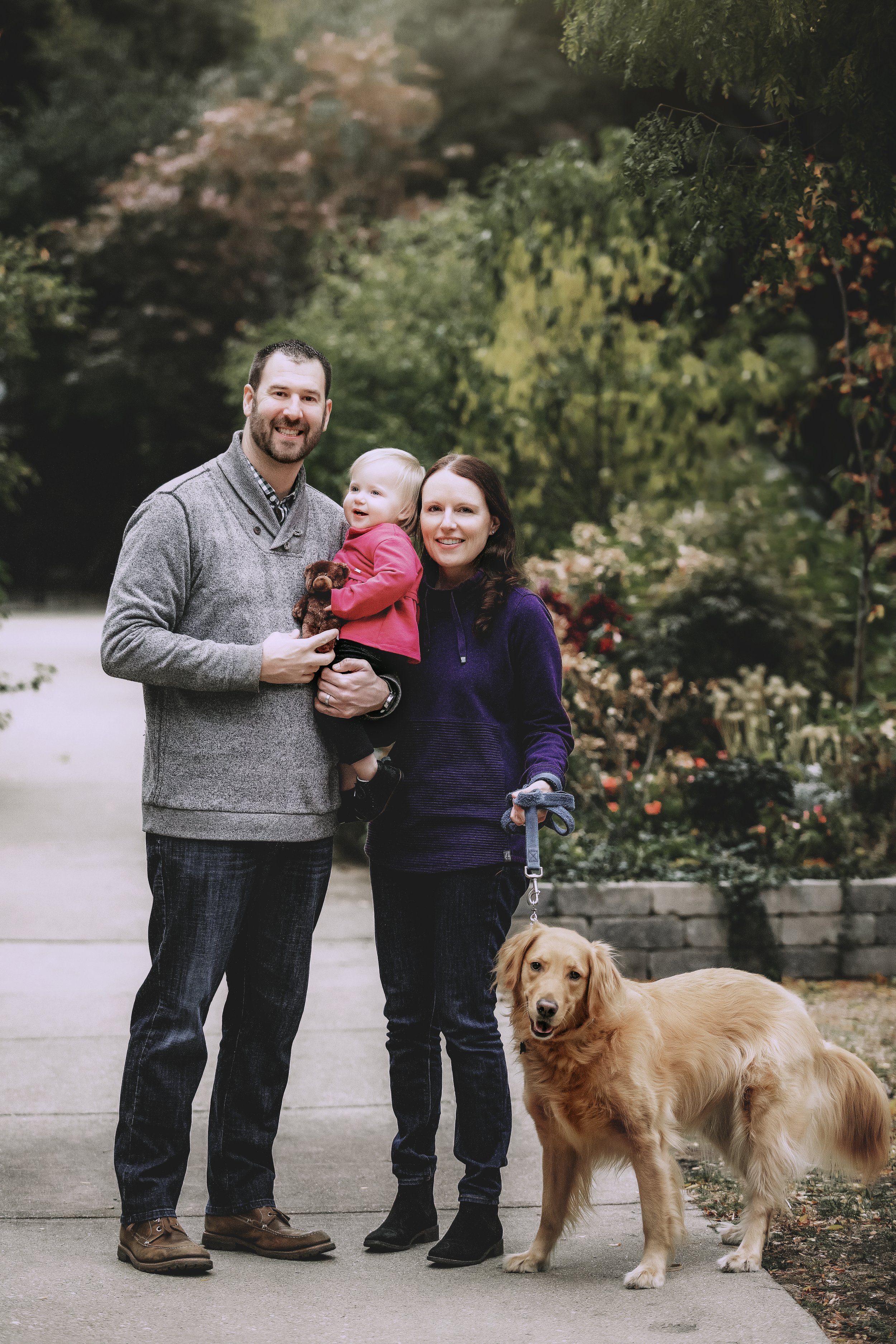 A family of three with a dog walking outdoors on a sidewalk, surrounded by trees and greenery.