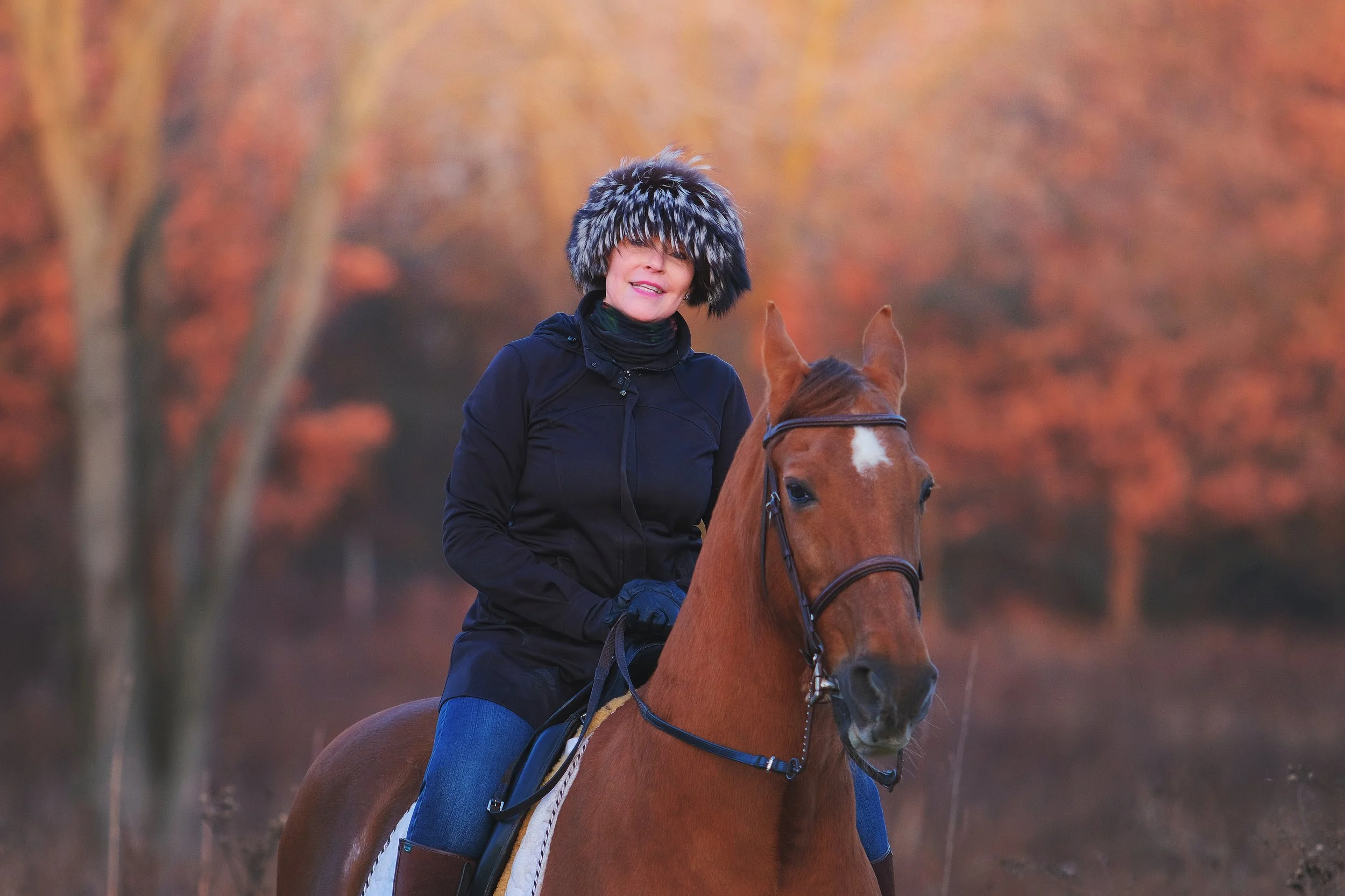 A woman riding a chestnut horse in a forest with autumn-colored leaves.