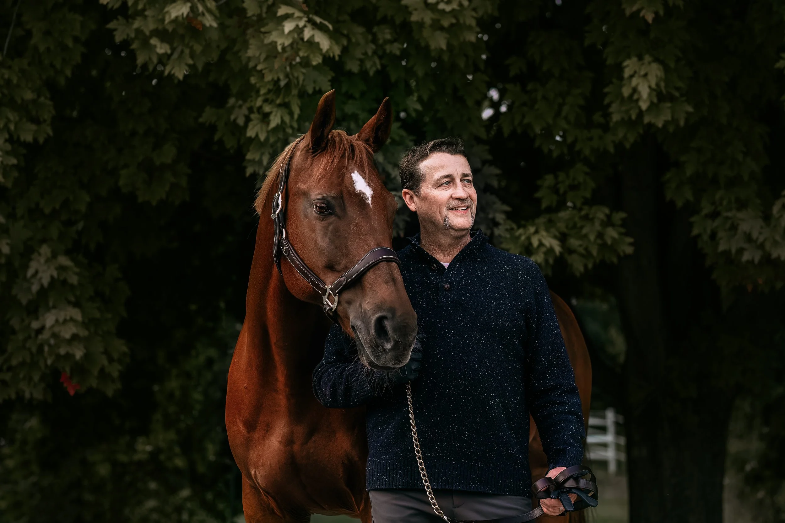A man standing beside a brown horse outdoors, with large green trees in the background.