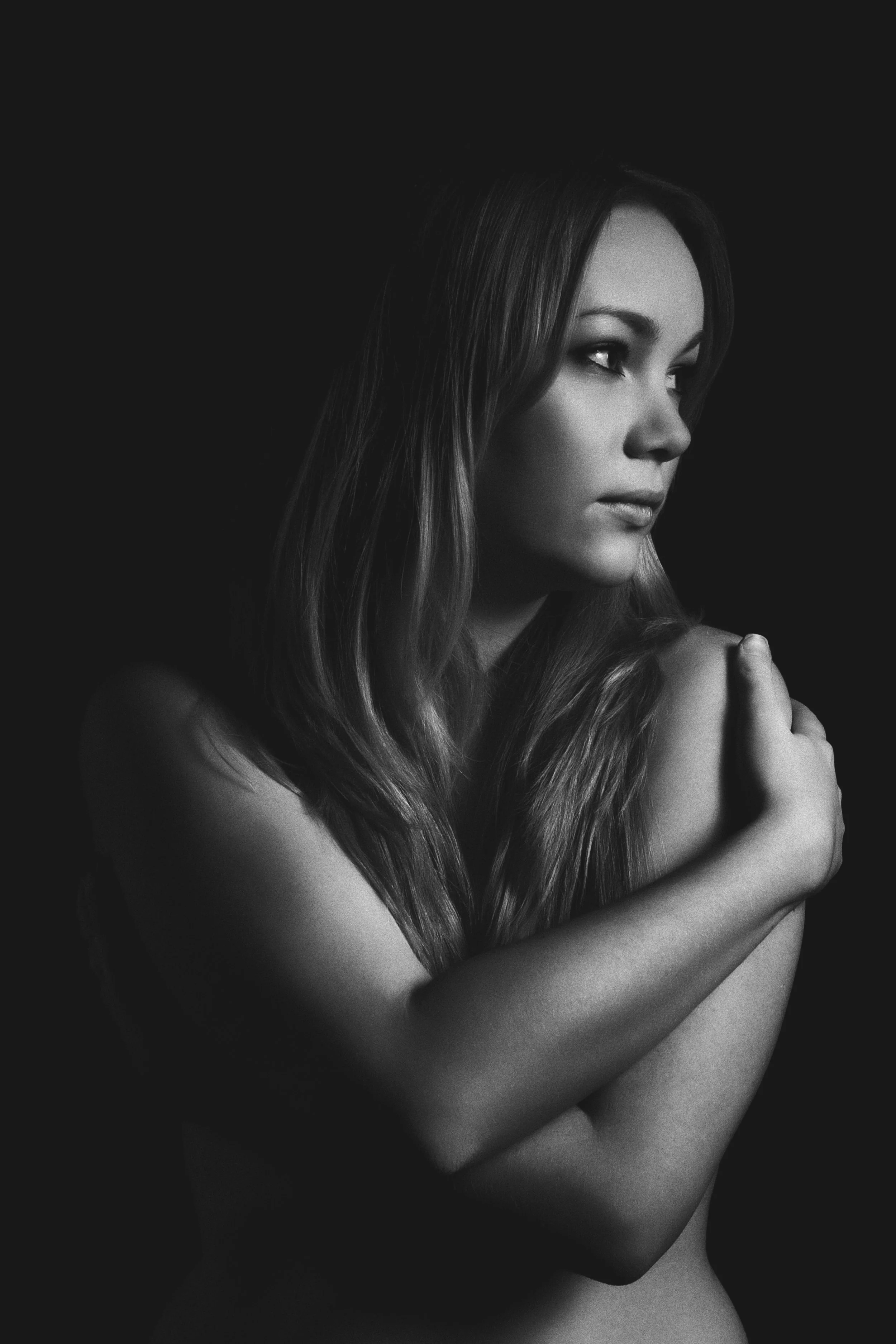 Black and white portrait of a woman with long hair, looking to her right, hugging her shoulder with her left hand, against a dark background.