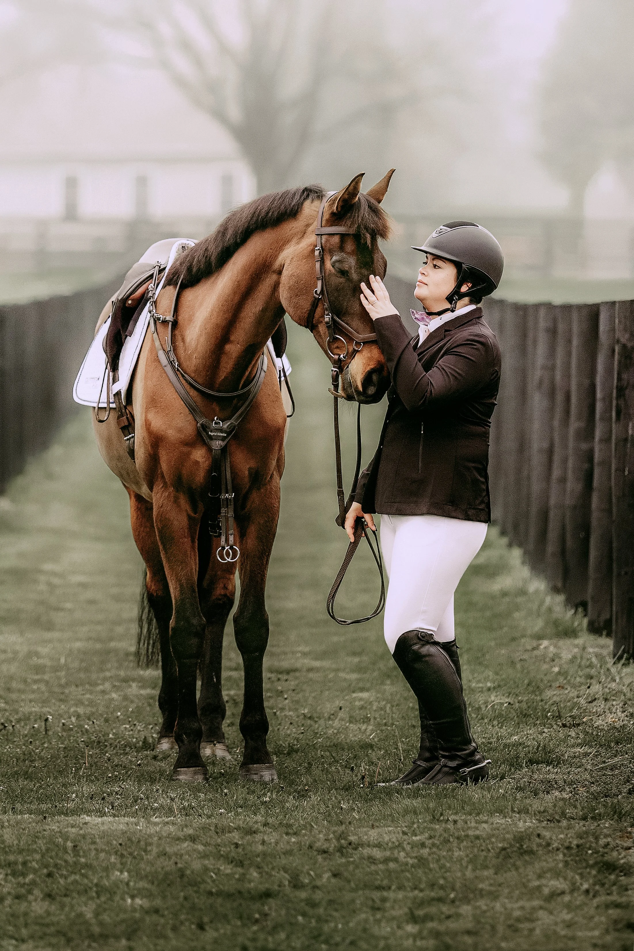 A woman dressed in equestrian attire, including white breeches and a black riding jacket, stands on a grassy area touching a brown horse's forehead. The horse is equipped with a saddle and bridle. The background is foggy with a wooden fence and a blu