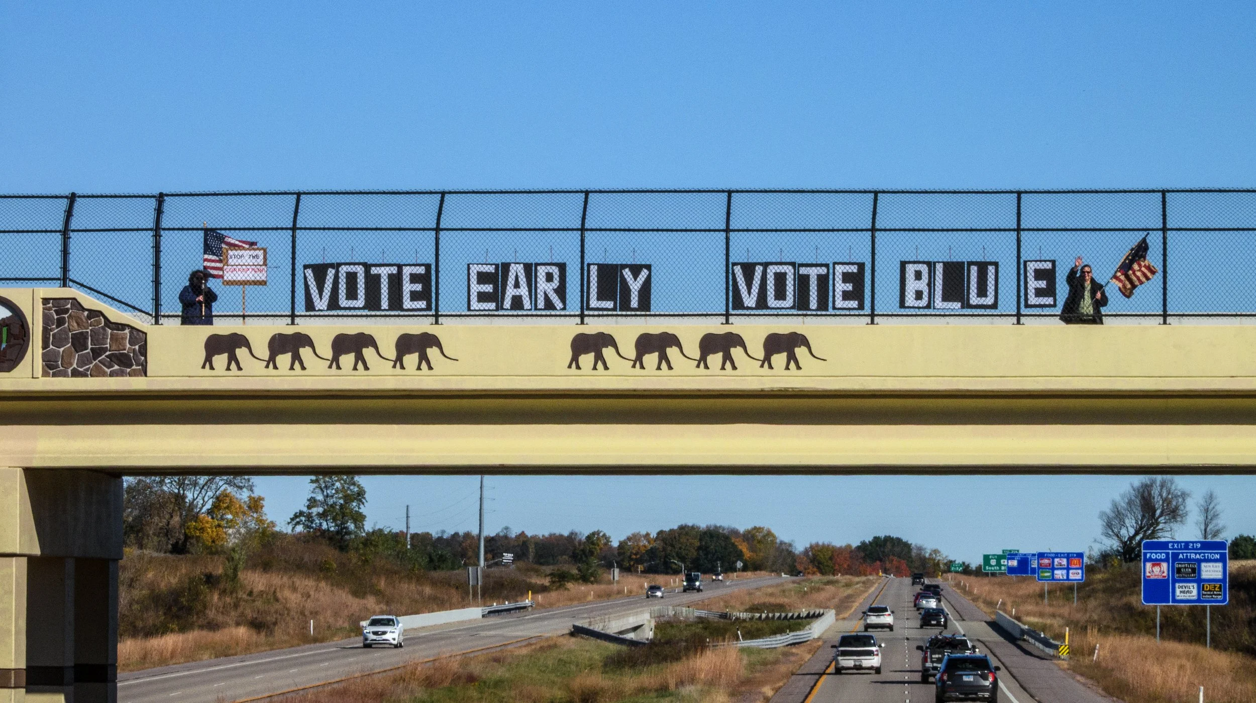 Two people stand on a bridge overpassing Rt. 12, having put up signs that read Vote Early, Vote Blue