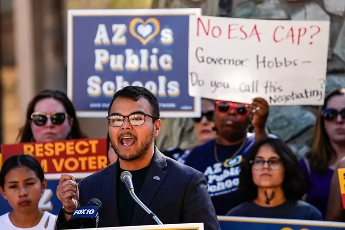 A man in glasses and a dark jacket speaking at a microphone during a protest, with signs behind him including one that reads 'AZ Public Schools' and another asking if there is no ESA cap with 'Governor Hobbs'.