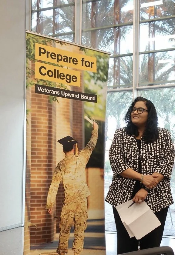 A woman with long dark hair and glasses standing next to a tall promotional banner. The banner has text that says 'Prepare for College' and 'Veterans Upward Bound,' and features an image of a person in military uniform graduation attire, holding a di