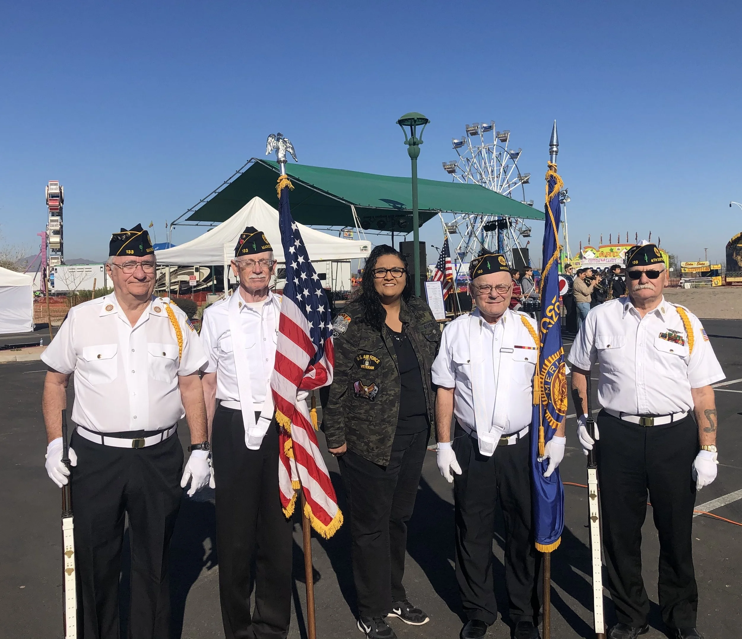 Group of five veterans standing together at an outdoor event, holding American flags, with amusement park rides in the background.