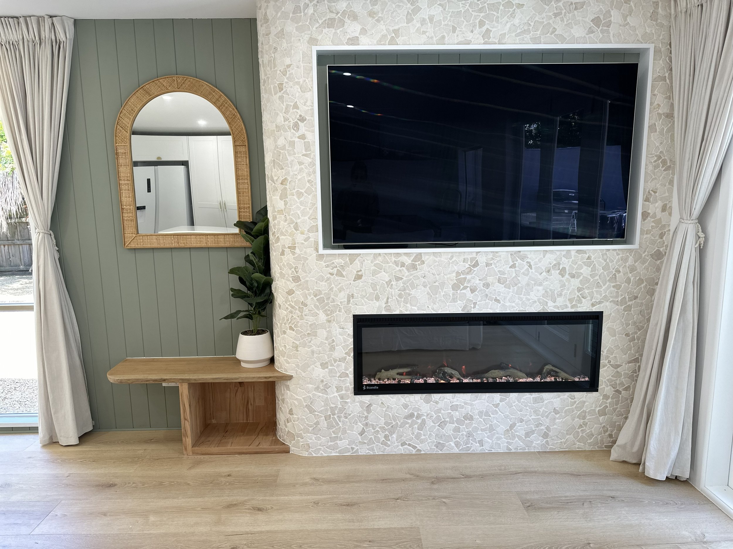 Living room with a flat-screen TV mounted on an off-white textured wall, a modern fireplace below the TV, a wooden bench with a potted plant, a mirror with a rattan frame, and curtains on both sides of the window.