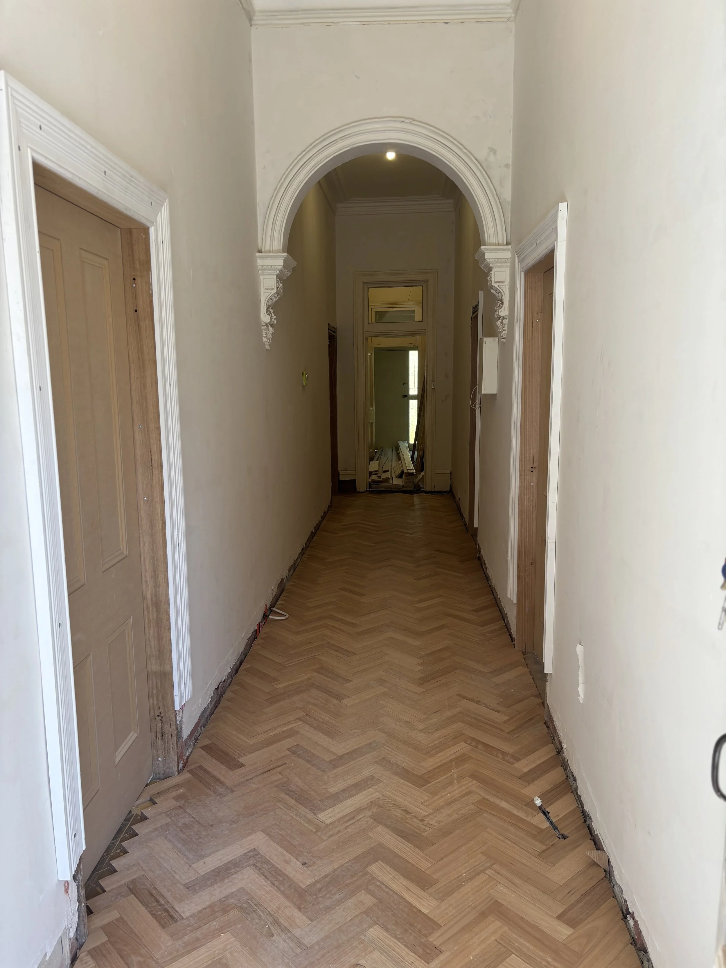 Interior view of a hallway with wooden chevron-patterned flooring, cream-colored walls, and decorative archways under construction.