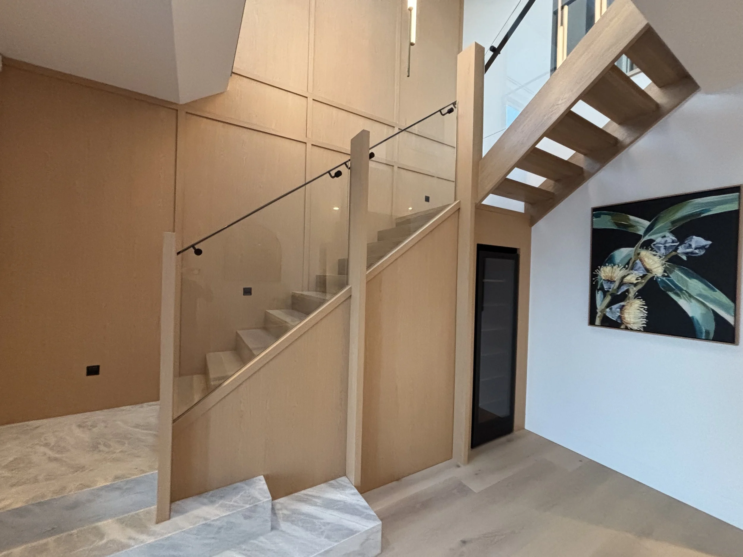 Interior view of a modern staircase with wooden steps, glass railing, and black hardware, next to a white wall with a colorful botanical painting.