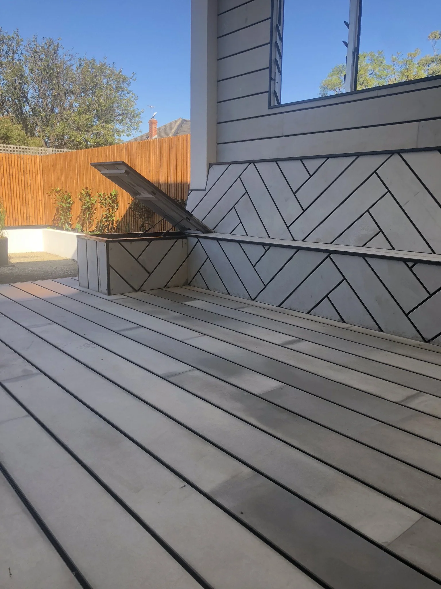 Outdoor patio with wooden decking, a built-in bench with a tiled exterior, a landscaped yard with small plants, a wooden fence, and a house wall with siding and a window.