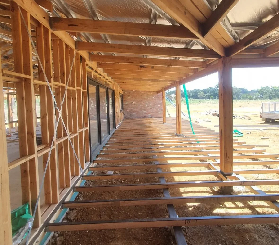 Construction site of a building with wooden framing, a brick wall in the background, unfinished flooring, and exposed roof beams, with construction equipment and open land outside.