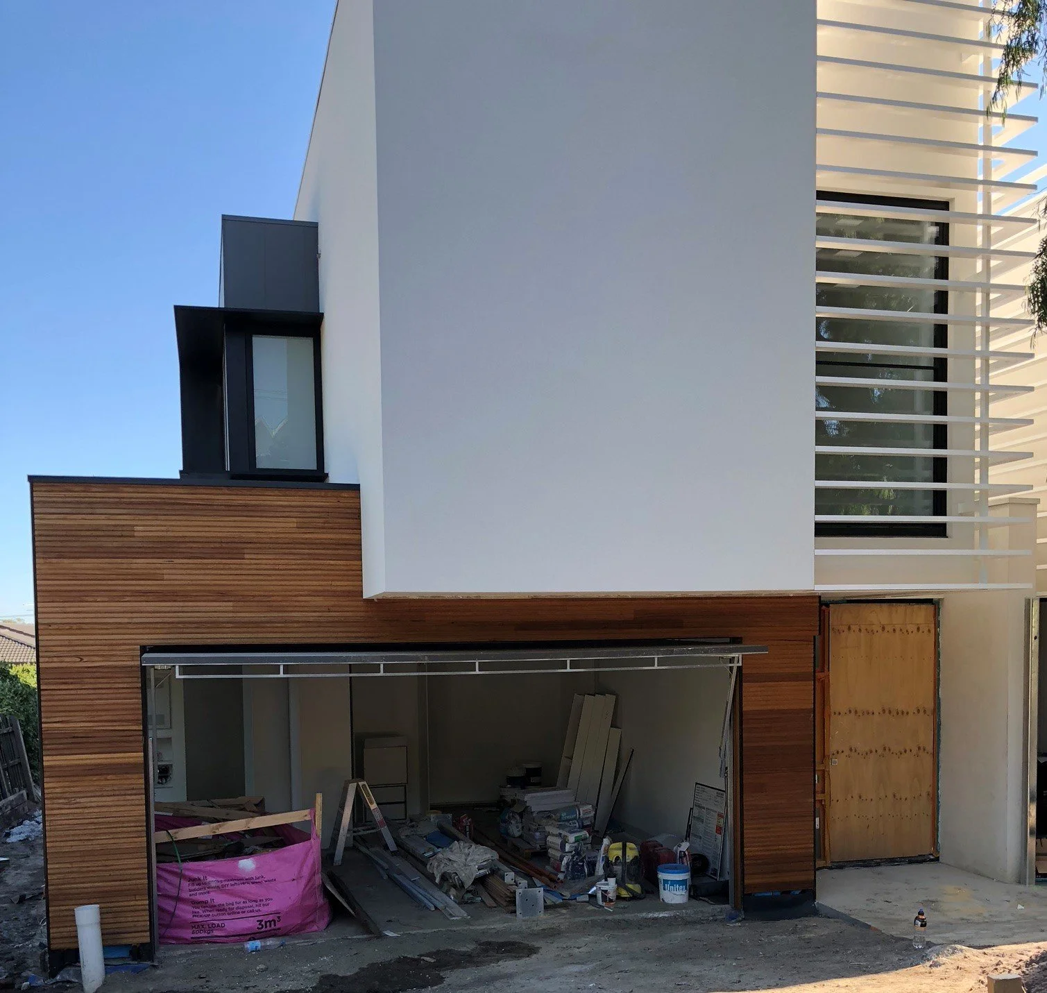 Modern multi-story house under construction with a white and wooden exterior, large window, and partially installed garage door, construction tools and materials inside.