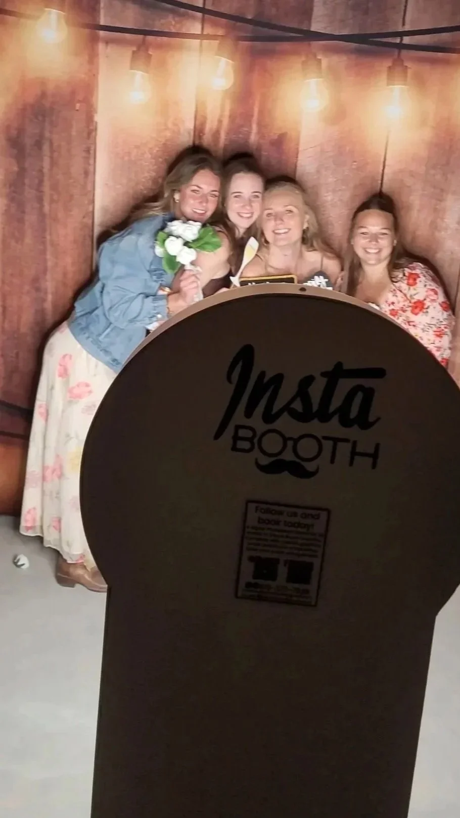 Four women smiling and posing for a photo in a photo booth with a wooden backdrop and string lights. The photo booth sign labeled 'Insta Booth' is visible in the foreground, partially covering the women.
