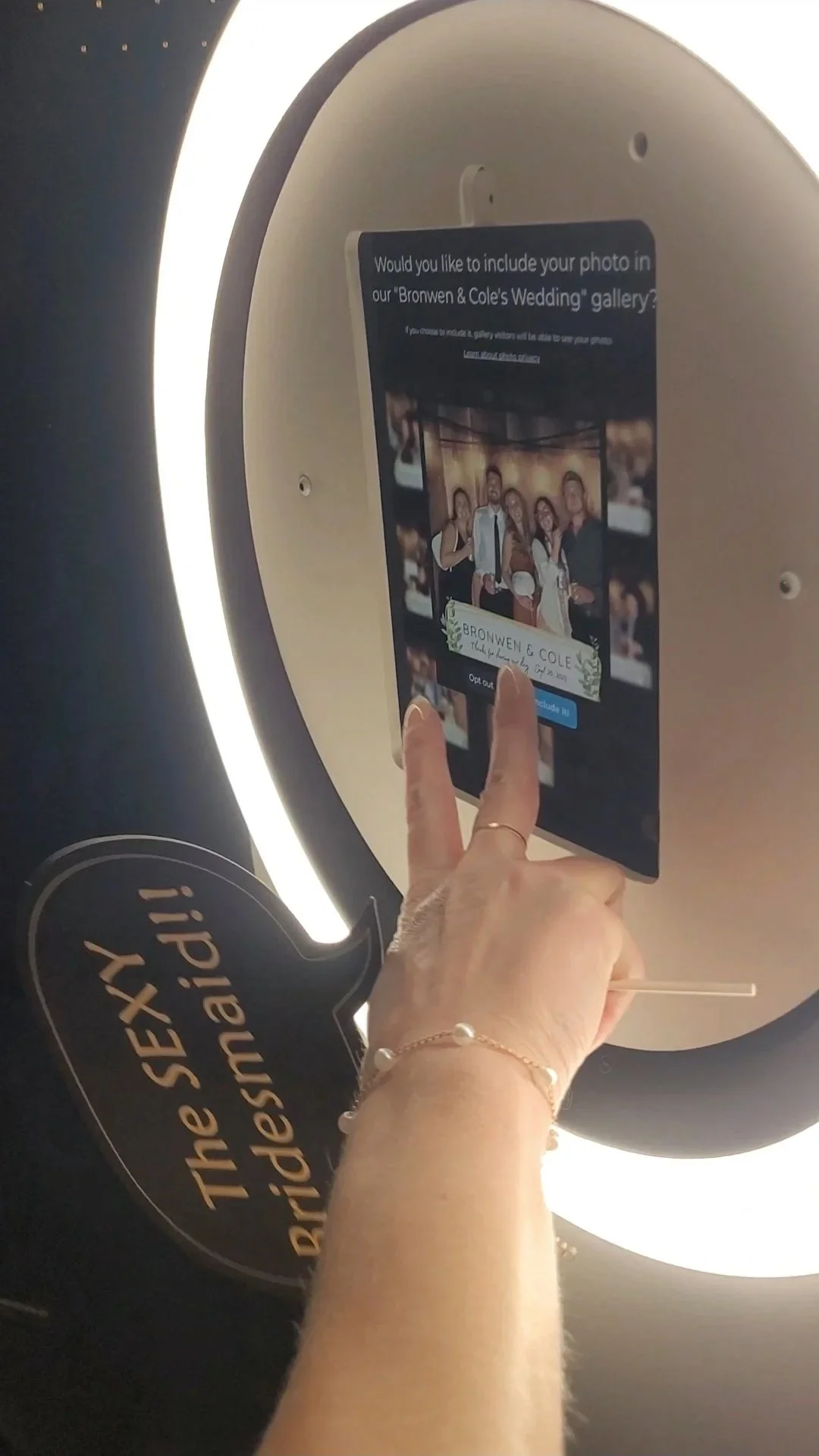 A person interacts with a photo kiosk, selecting a wedding photo to include in a digital gallery labeled 'Bronwen & Cole's Wedding'.