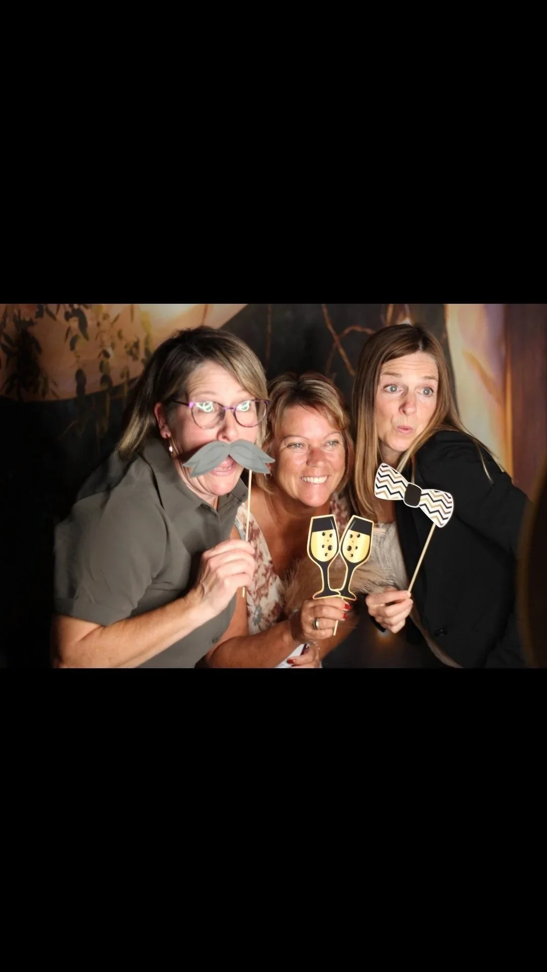Three women at a celebration, holding photo booth props including a mustache, champagne glasses, and a bow tie, smiling and making playful faces.