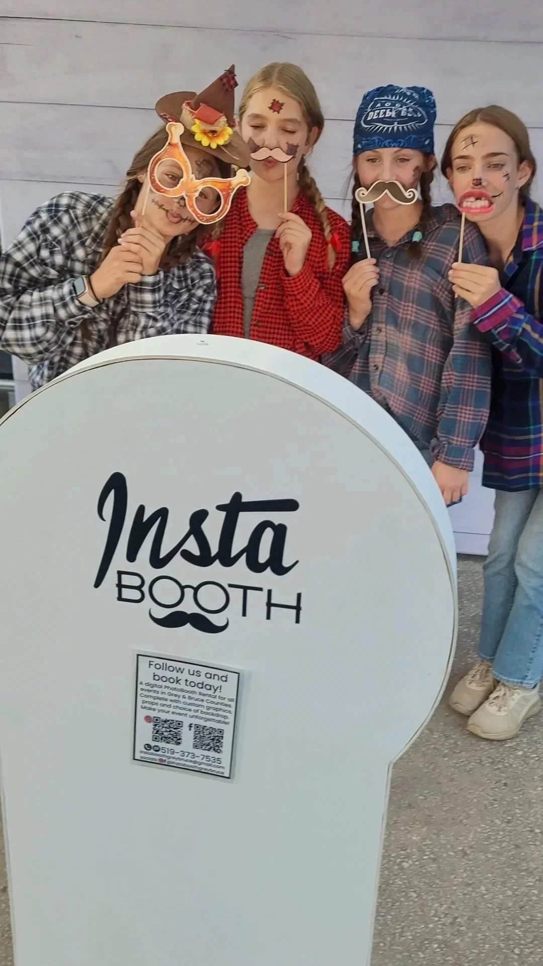 Four girls in plaid shirts holding photo booth props, standing behind an 'Insta Booth' sign with a QR code and contact information.