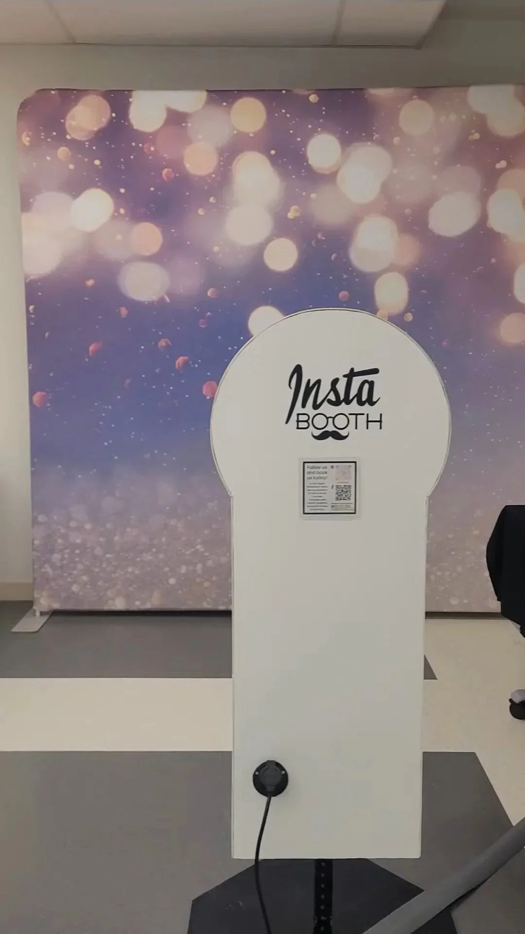 Photo of a photo booth labeled 'Insta Booth' with a background of purple and pink bokeh lights. There is a QR code on the side of the booth.