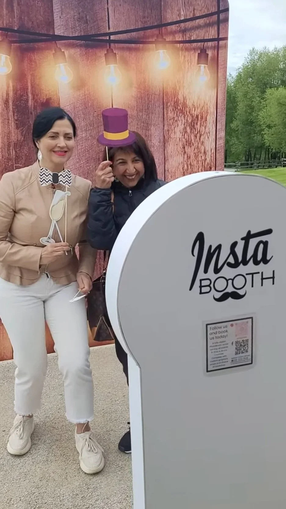 Two women at an Instagram photo booth with a rustic wood background, holding photo props and wearing a paper hat, smiling for the camera.