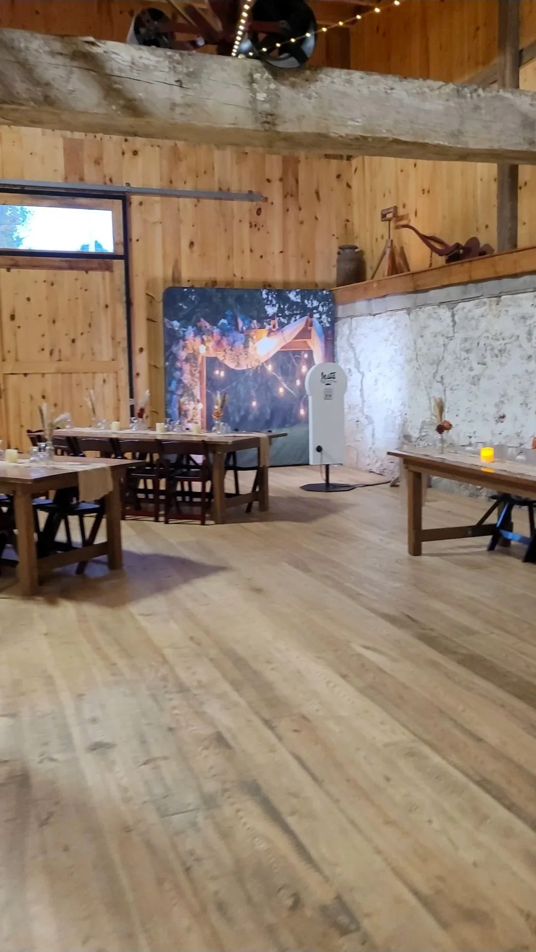 Rustic indoor space with wooden walls and flooring, decorated with table settings and fairy lights, featuring a large photo backdrop and a small vintage photo booth, likely prepared for a celebration or event.