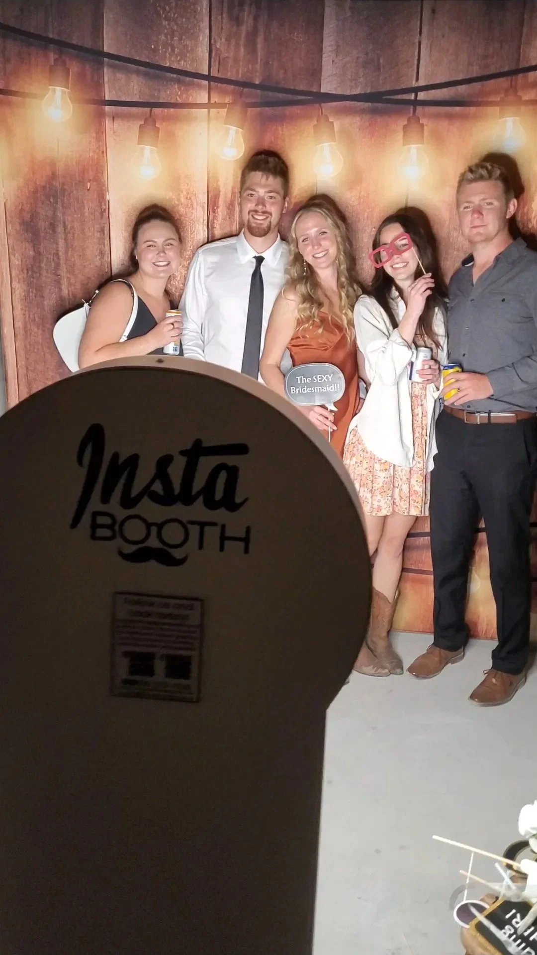 Group of five young adults at a photo booth, with a wooden background and hanging light bulbs. Two women are holding props, one with red glasses and the other with a speech bubble that says "The SEXY Bridesmaid!!". The group looks happy and is dresse
