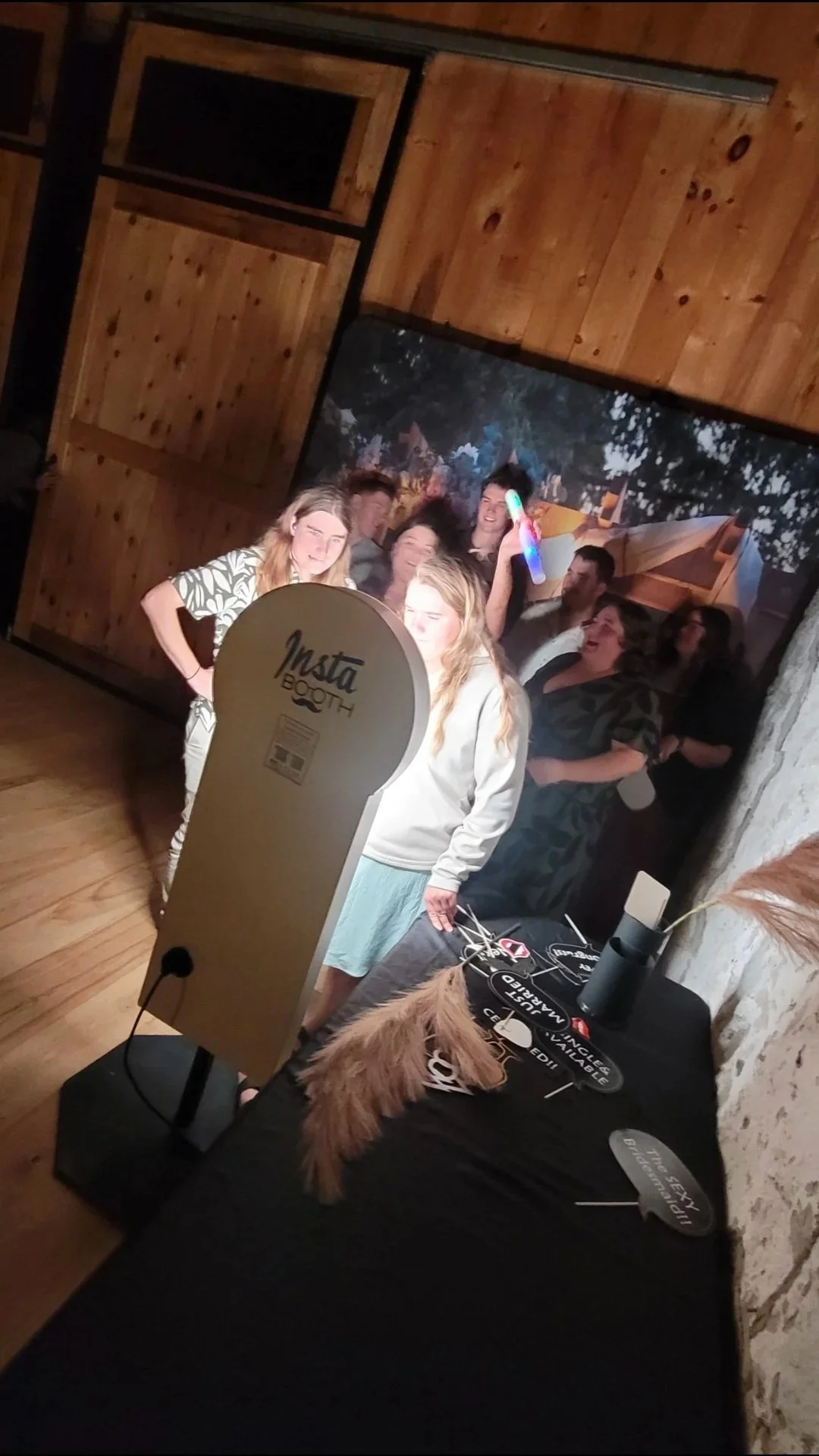 A group of people at a party taking a photo at an Insta booth, with one person holding a glow stick, inside a rustic wooden room with a large mural or photograph of a tent scene in the background.