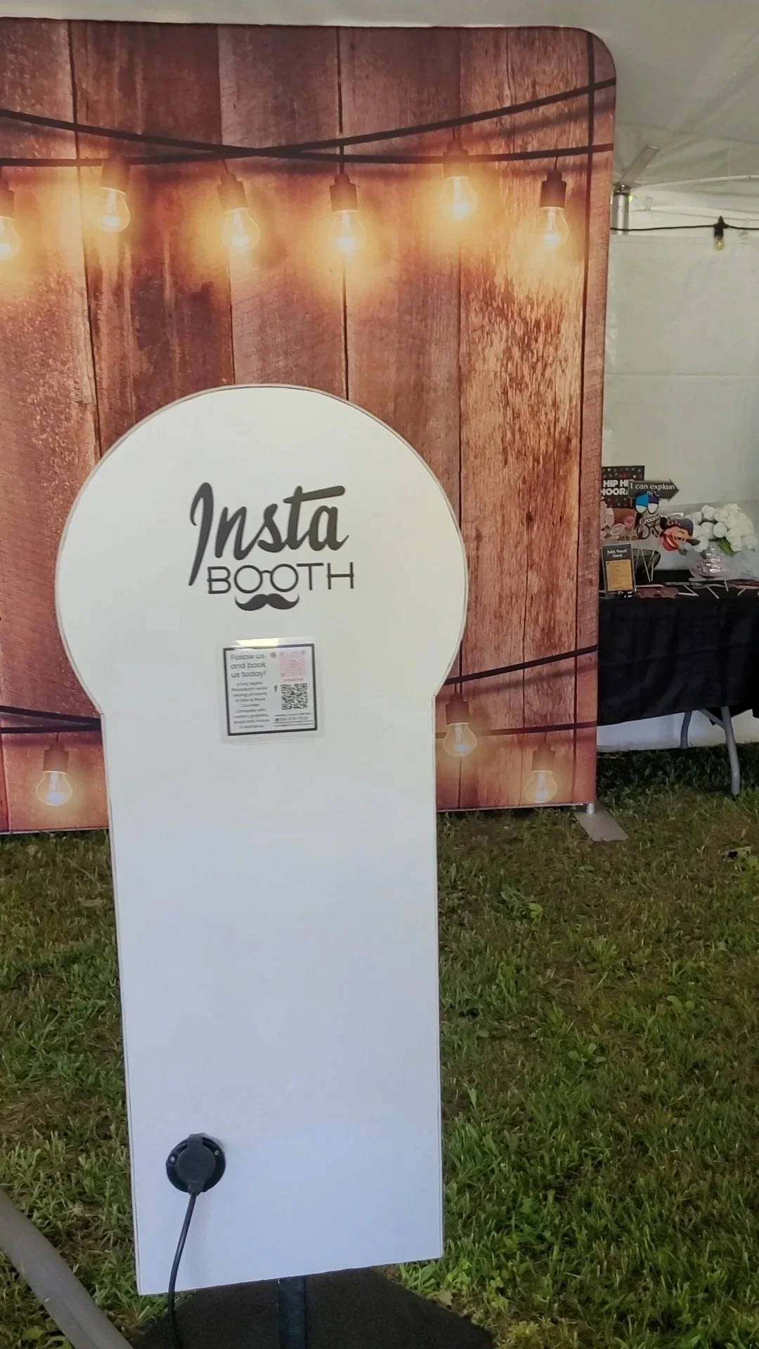 Sign for an Instagram photo booth at an event, with a wooden background and string lights above, on a grassy area.
