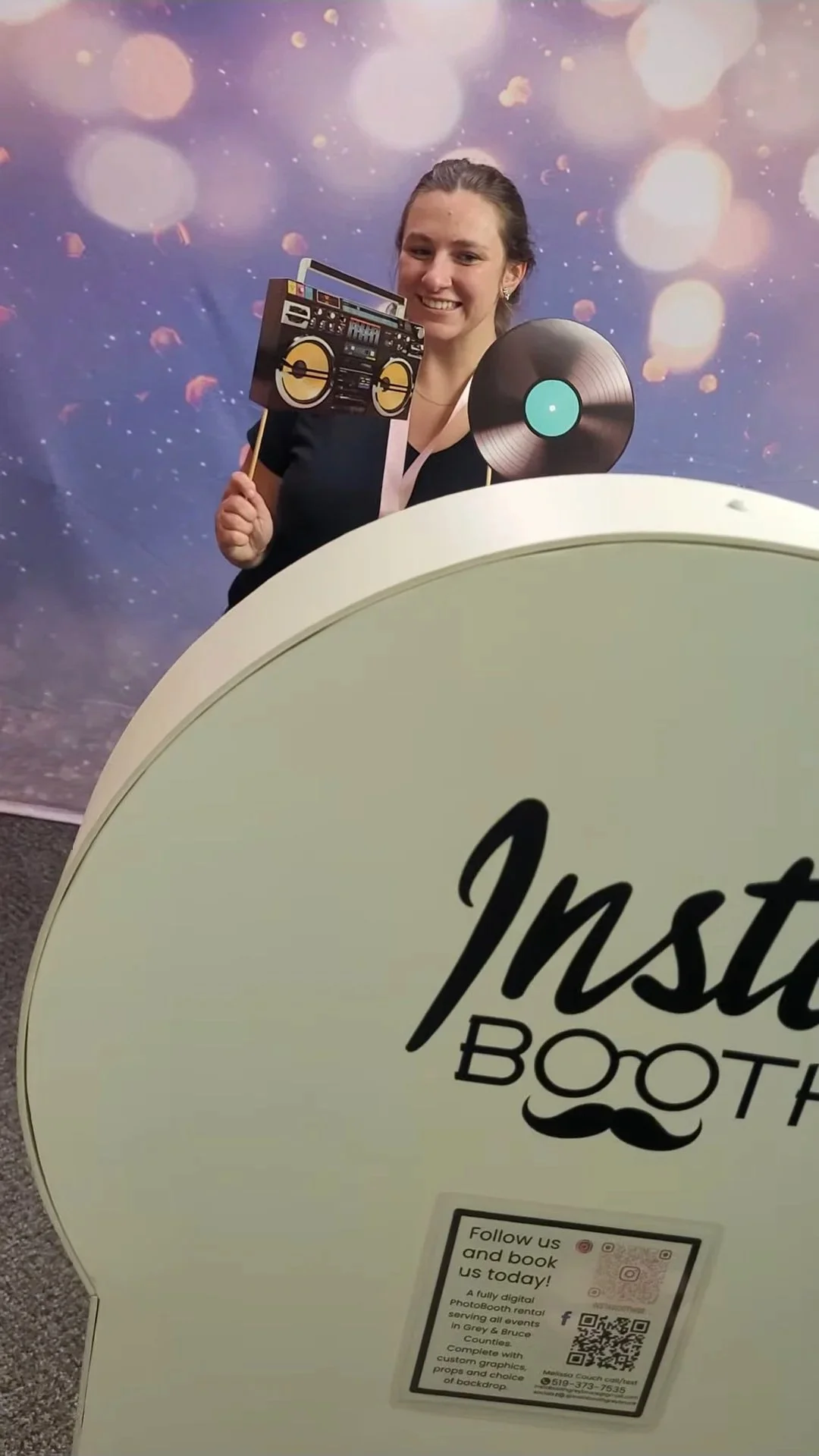 Woman smiling in a photo booth holding a photo prop of a vinyl record and turntable, with a backdrop with bokeh light effects, in front of a sign that says 'Instant Booth'.