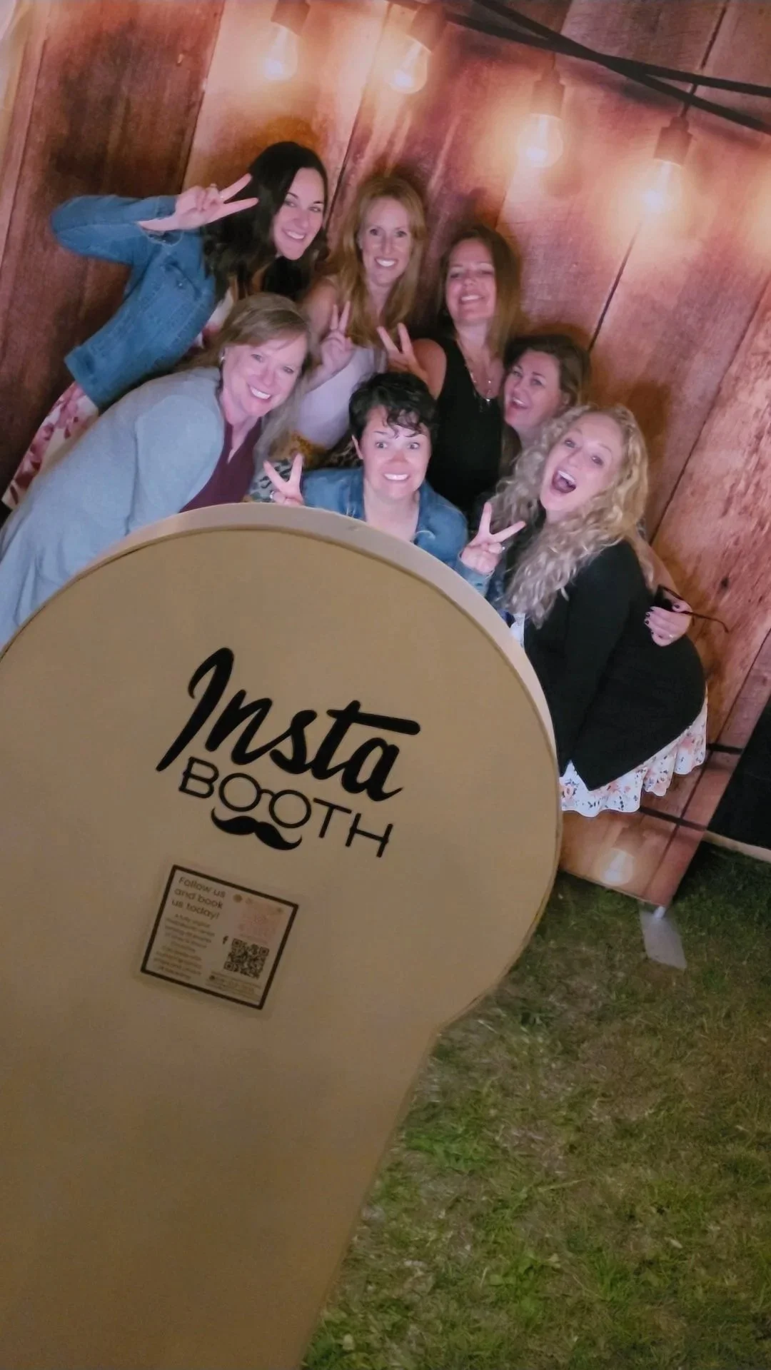 A group of eight women posing together at an Insta Booth, making peace signs and smiling. The photo is taken at an event with a wooden background and string lights. The Insta Booth sign is visible in the foreground.