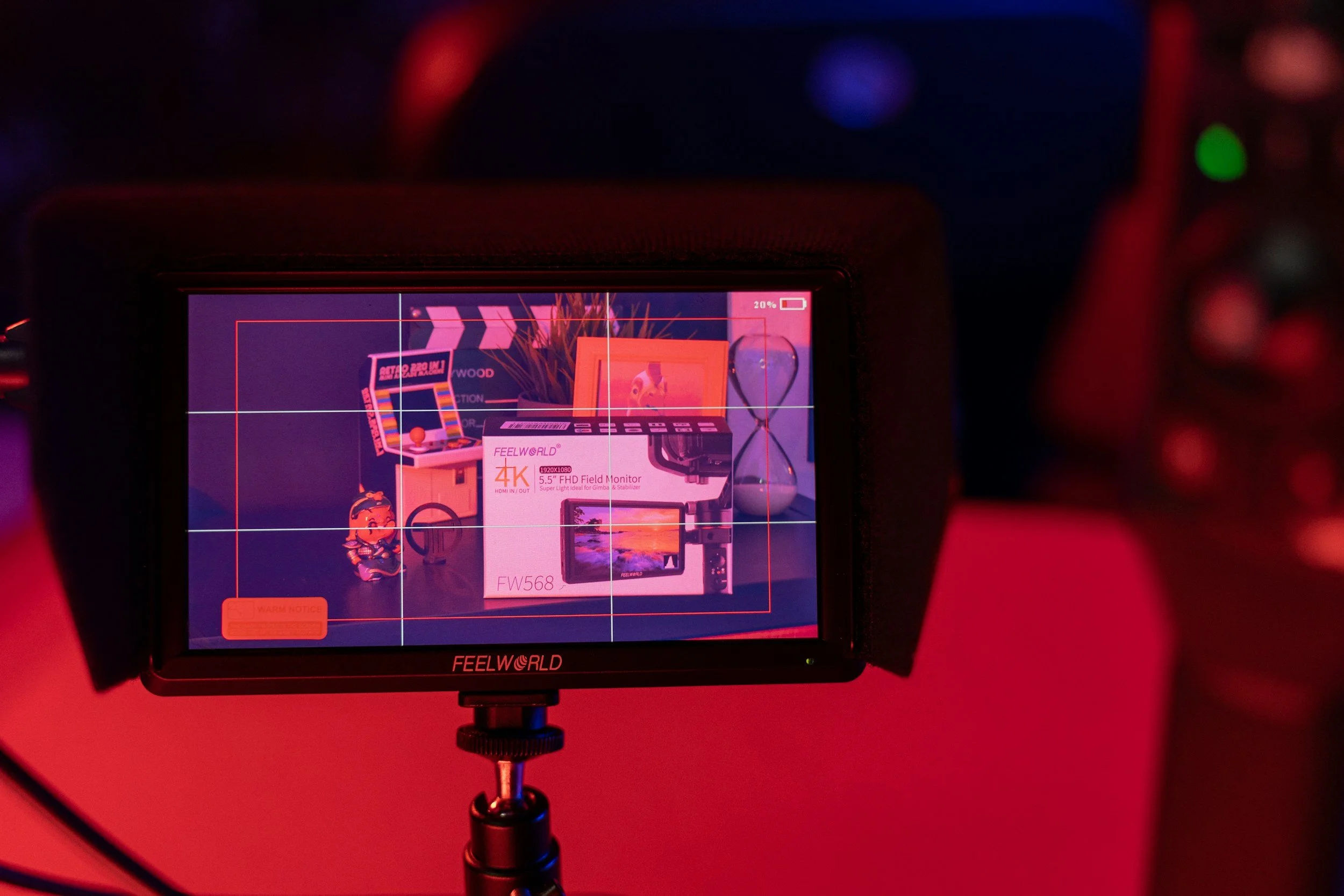 A camera screen displays a product shot of a Feelworld 4K 5.5-inch FHD field monitor box on a colorful surface with a hourglass, a framed photo, and small objects, with red and blue lighting.