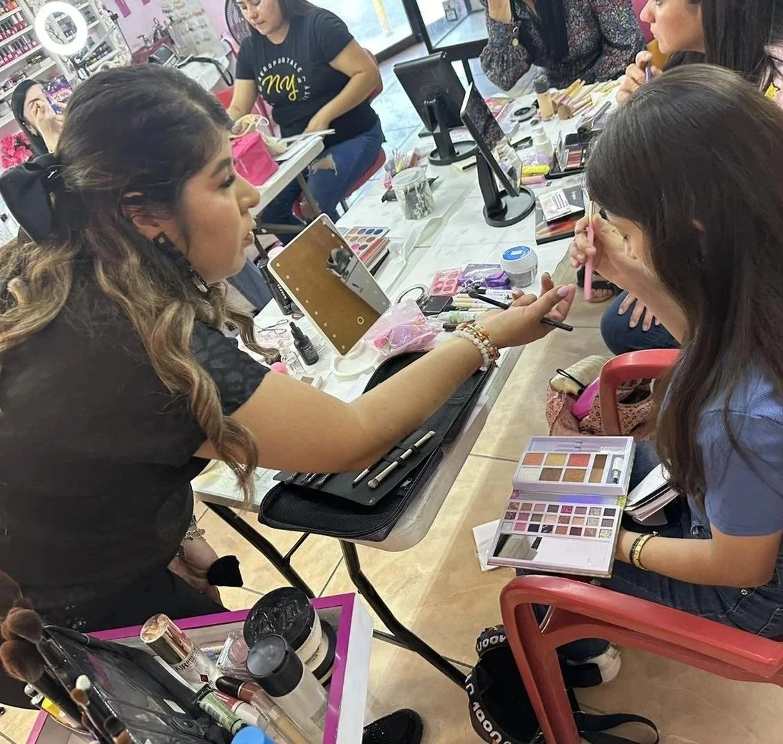 Multiple women gathered around a table participating in a makeup class or tutorial, with eyeshadow palettes, makeup brushes, and tools spread out on the table.