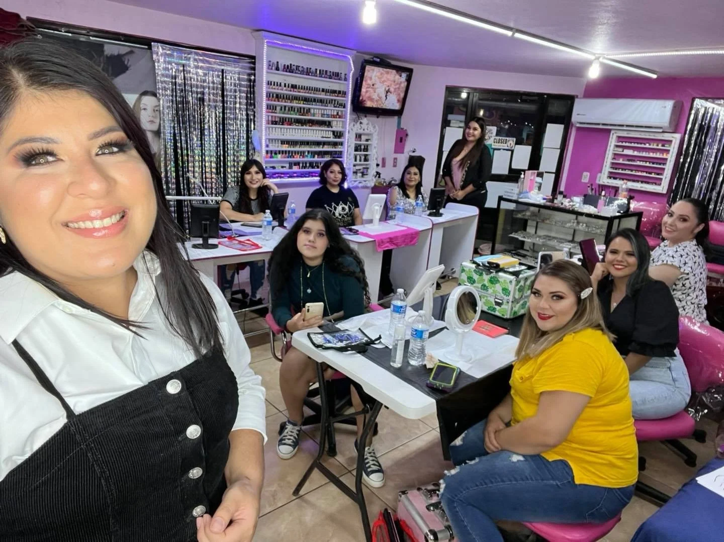 Group of women in a nail salon, some sitting at tables with nail polish displays in the background, getting nails done and smiling at the camera.