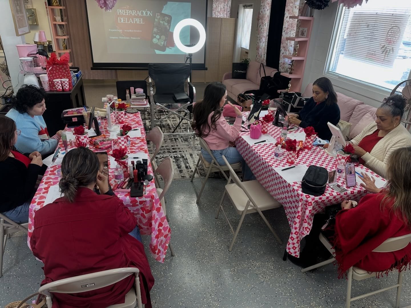 A group of women gathered around two long tables with pink and red checkered tablecloths, decorated with red roses, in a room set for a makeup or beauty workshop, with a présentation projected on a screen