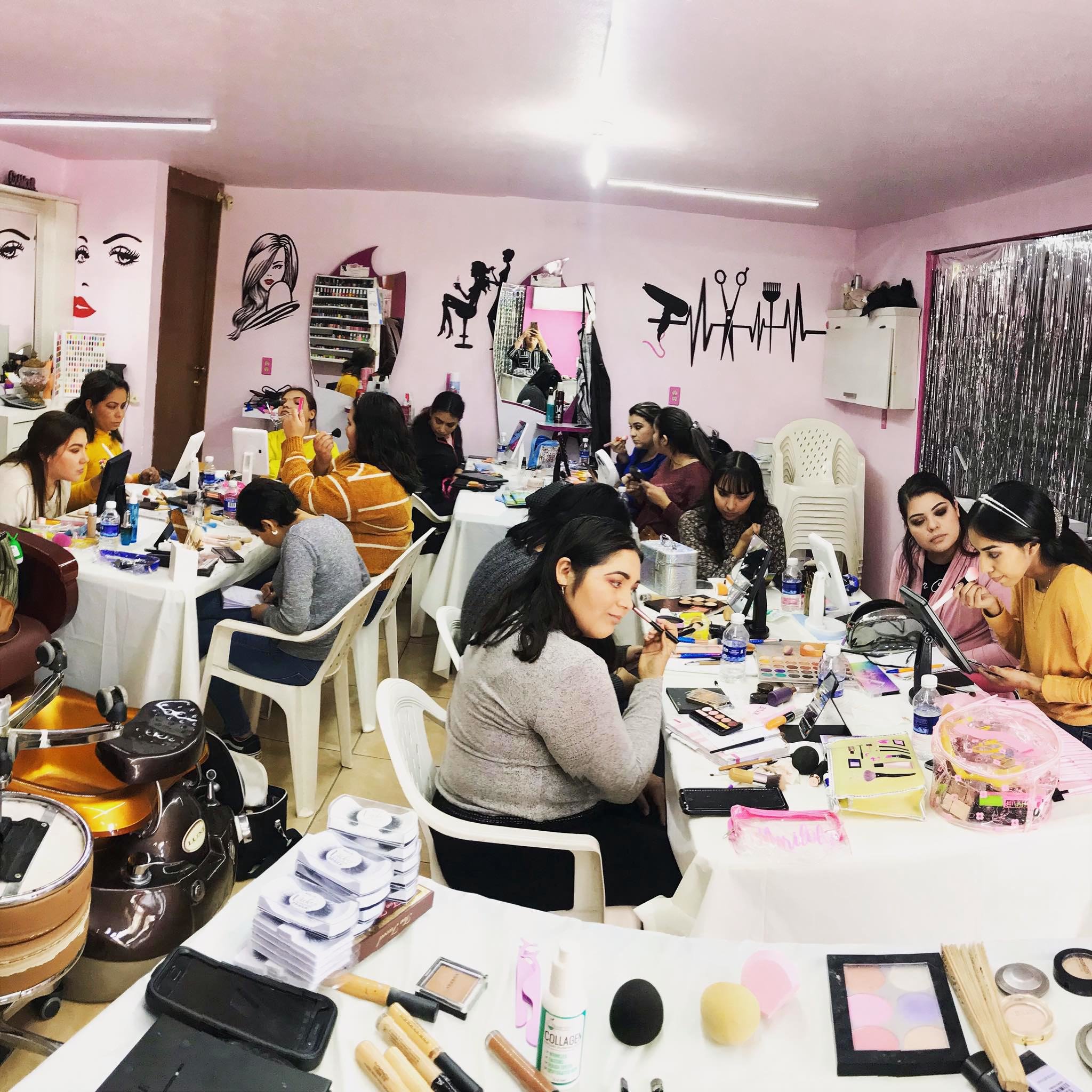 Group of women sitting at tables applying makeup, with beauty products and tools on the tables, and wall decor related to beauty and makeup in the background.