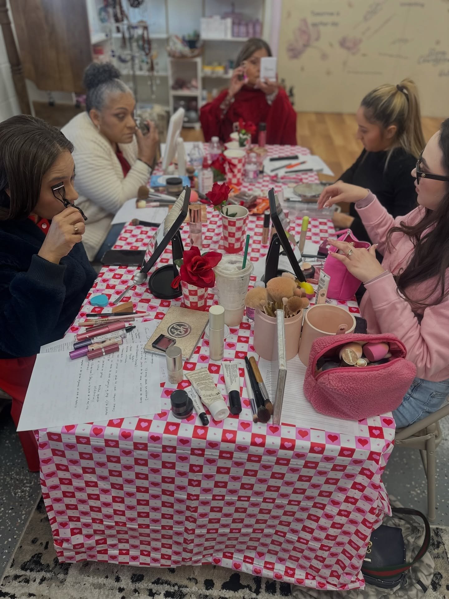Women participating in a makeup workshop around a table covered with a red and white checkered tablecloth, various makeup products, and handheld mirrors, in a room decorated with soft colors and wooden floors.