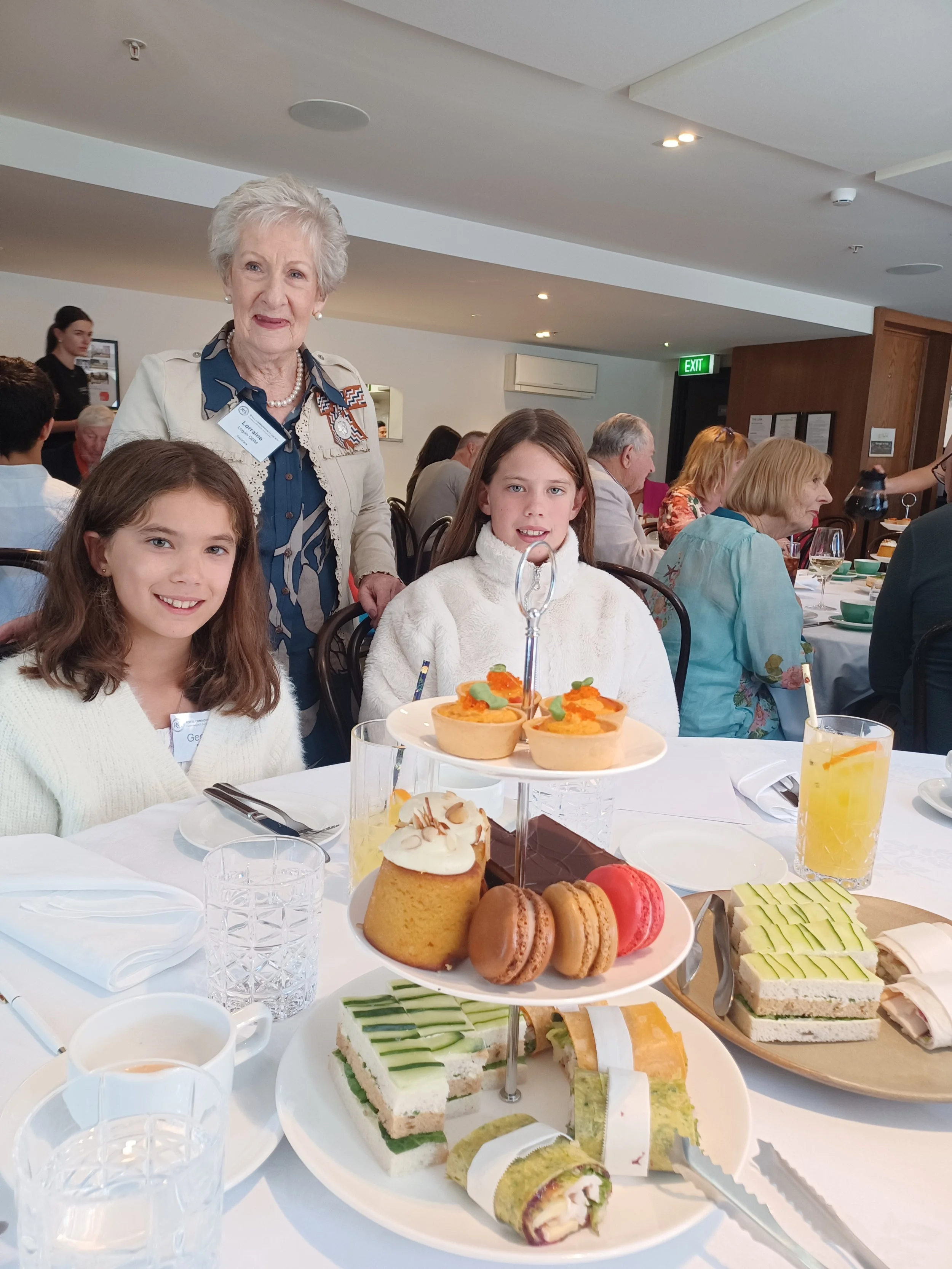 A table set for a meal with desserts and sandwiches, including cupcakes, macarons, layered cakes, and finger sandwiches, with people dining in the background.