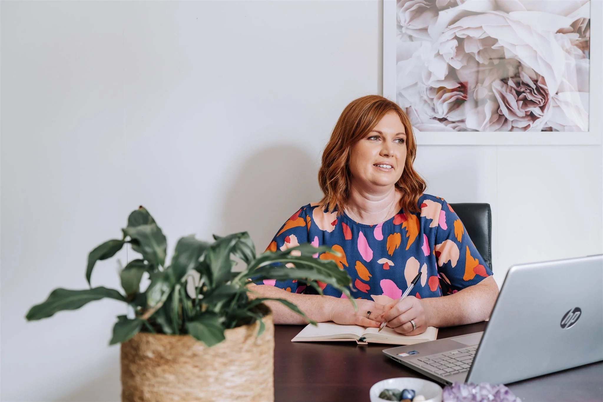 A woman with red hair sitting at a desk with a laptop, notebook, and pen, in an office with a white wall and a floral picture in the background, and a plant in the foreground.