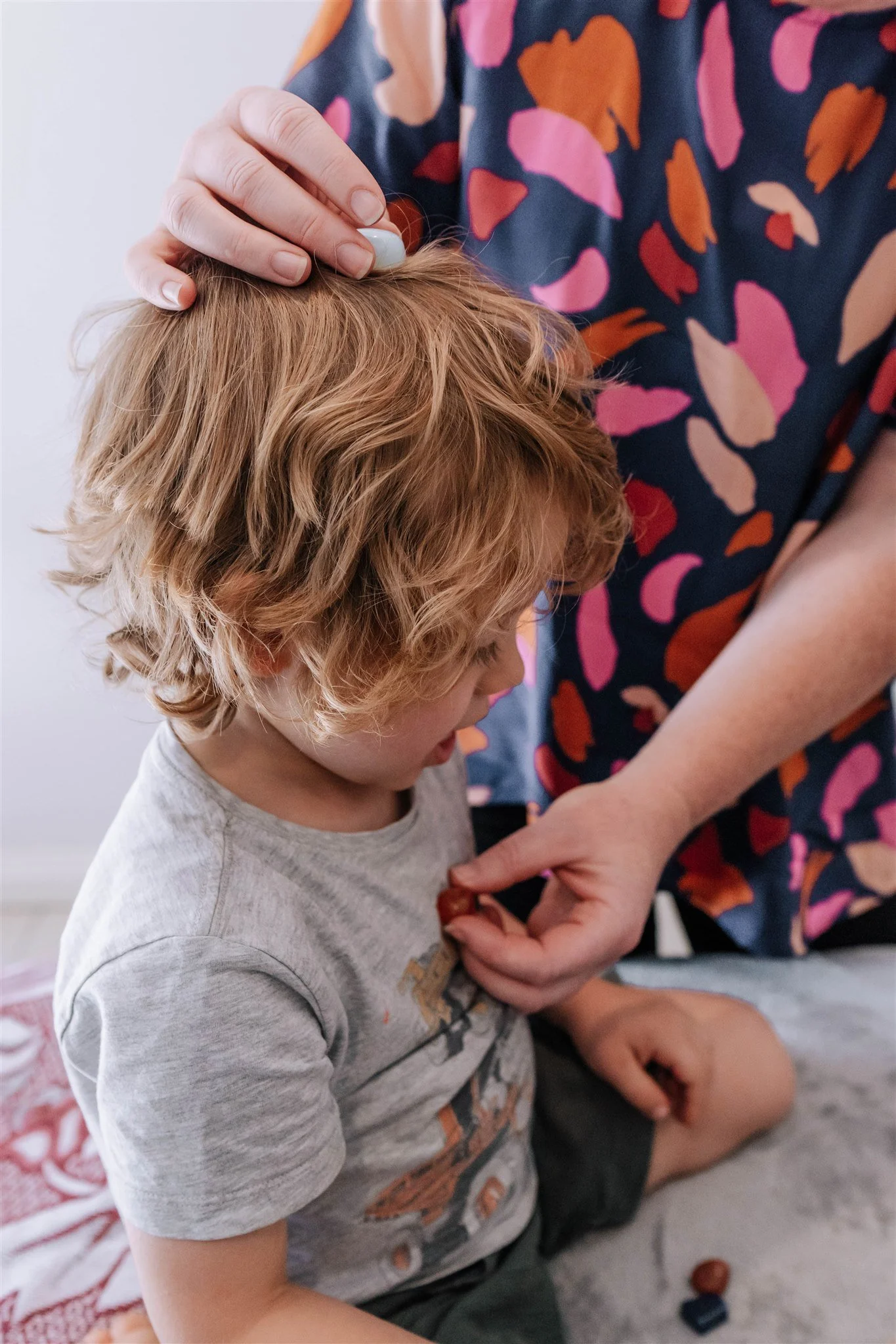 A child with curly red hair looking down at a chocolate treat being handed to him by an adult. The adult is holding the child’s head with one hand and the treat with the other. The child is sitting on a gray surface with some toys nearby. The adult wears a dark, colorful, patterned shirt.