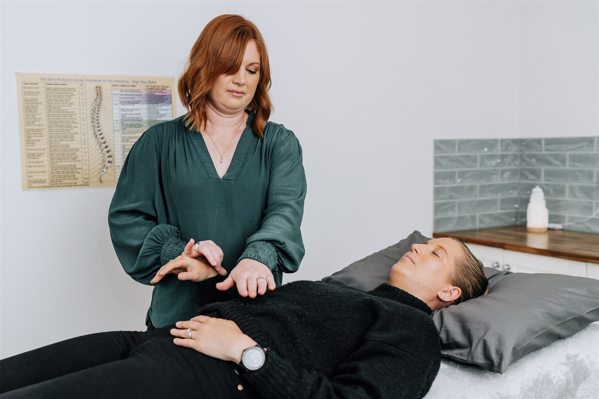 A woman in a green top is performing a medical examination or treatment on another woman who is lying on a bed with her eyes closed, in a clinical setting.