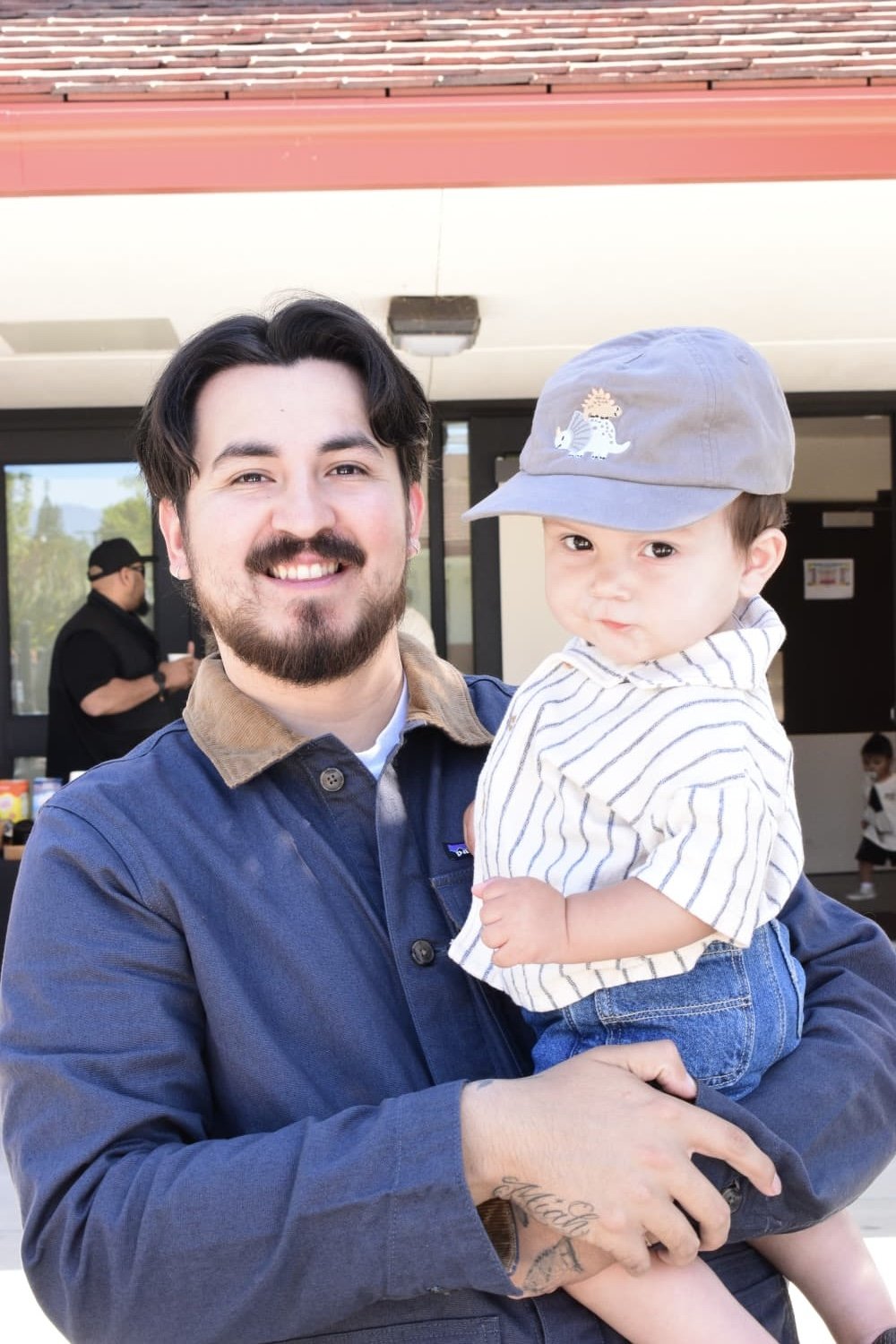A man holding a young boy outside a building, smiling at the camera. The boy wears a gray cap with a dinosaur and an animal embroidered on it, a striped shirt, and jeans.